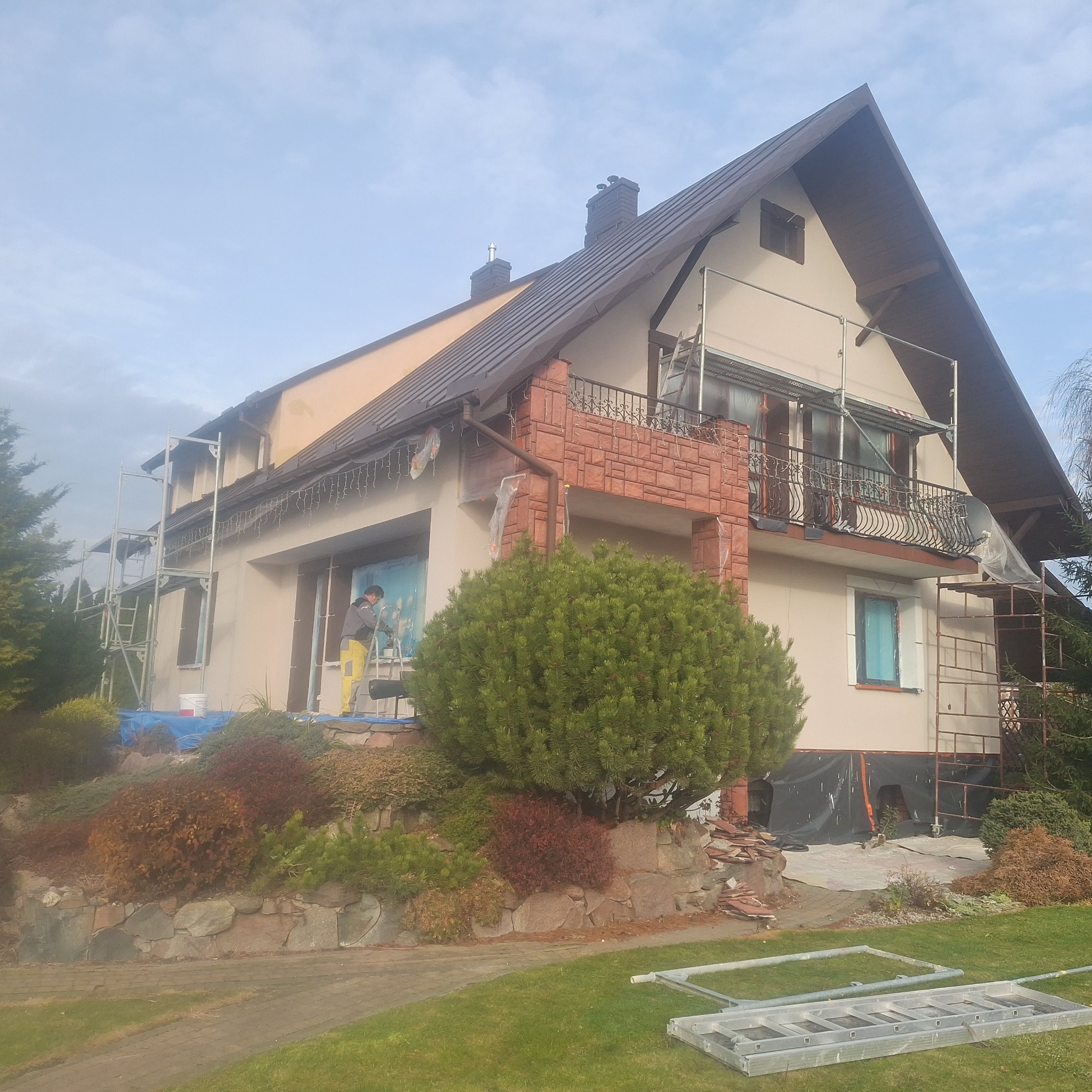 Dwupiętrowy dom z brązowym dachem w trakcie malowania elewacji. Widoczne rusztowania, zabezpieczenia okien folią i pracownik w żółtych spodniach.