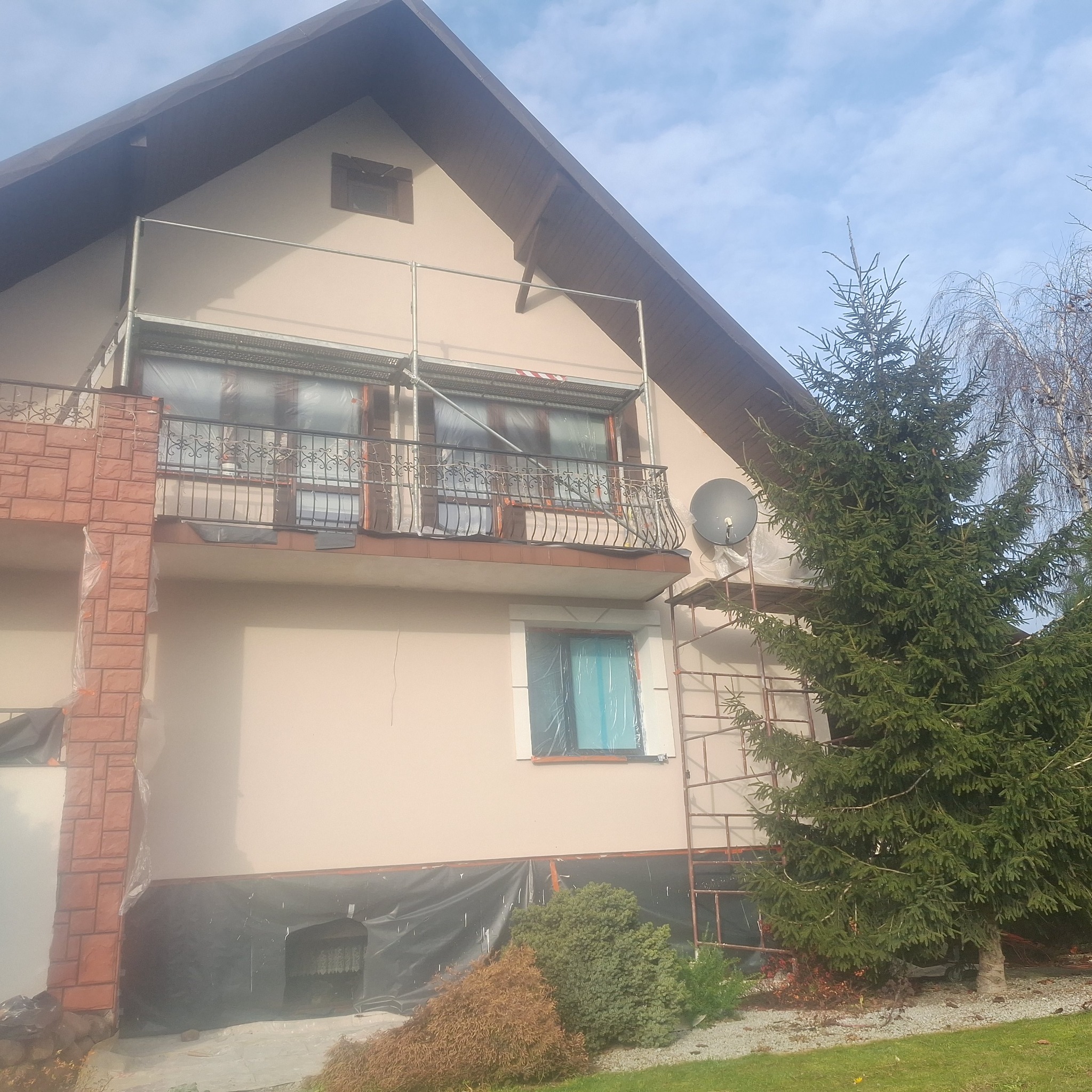 Dom jednorodzinny w trakcie prac elewacyjnych: zabezpieczone okna folią, rusztowania, świeżo pomalowana fasada w odcieniach beżu. Widok z poziomu gruntu.