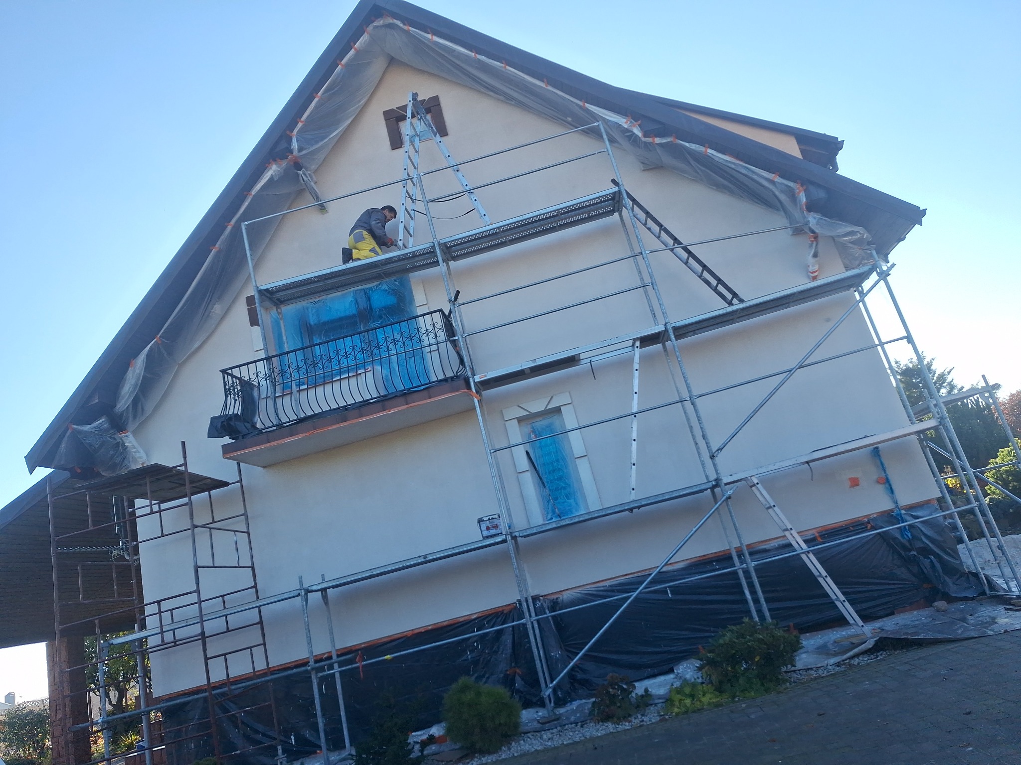Pracownik na rusztowaniu, zabezpieczony dom folią, przygotowuje elewację do malowania. Widoczny fragment dachu i okna zabezpieczone folią. Ujęcie z dołu.