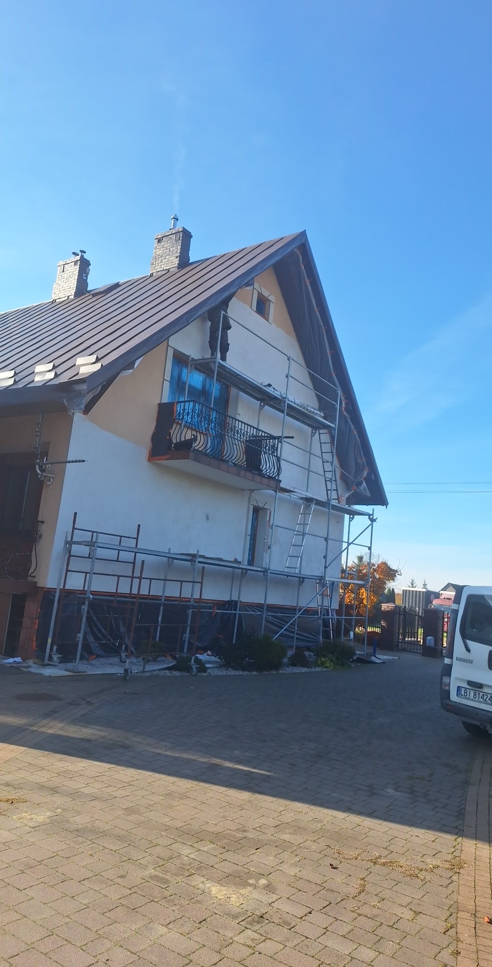 Dom jednorodzinny w trakcie malowania elewacji, z widocznymi rusztowaniami i zabezpieczonym balkonem. Część ściany pomalowana na jasny kolor, reszta przygotowana do malowania.