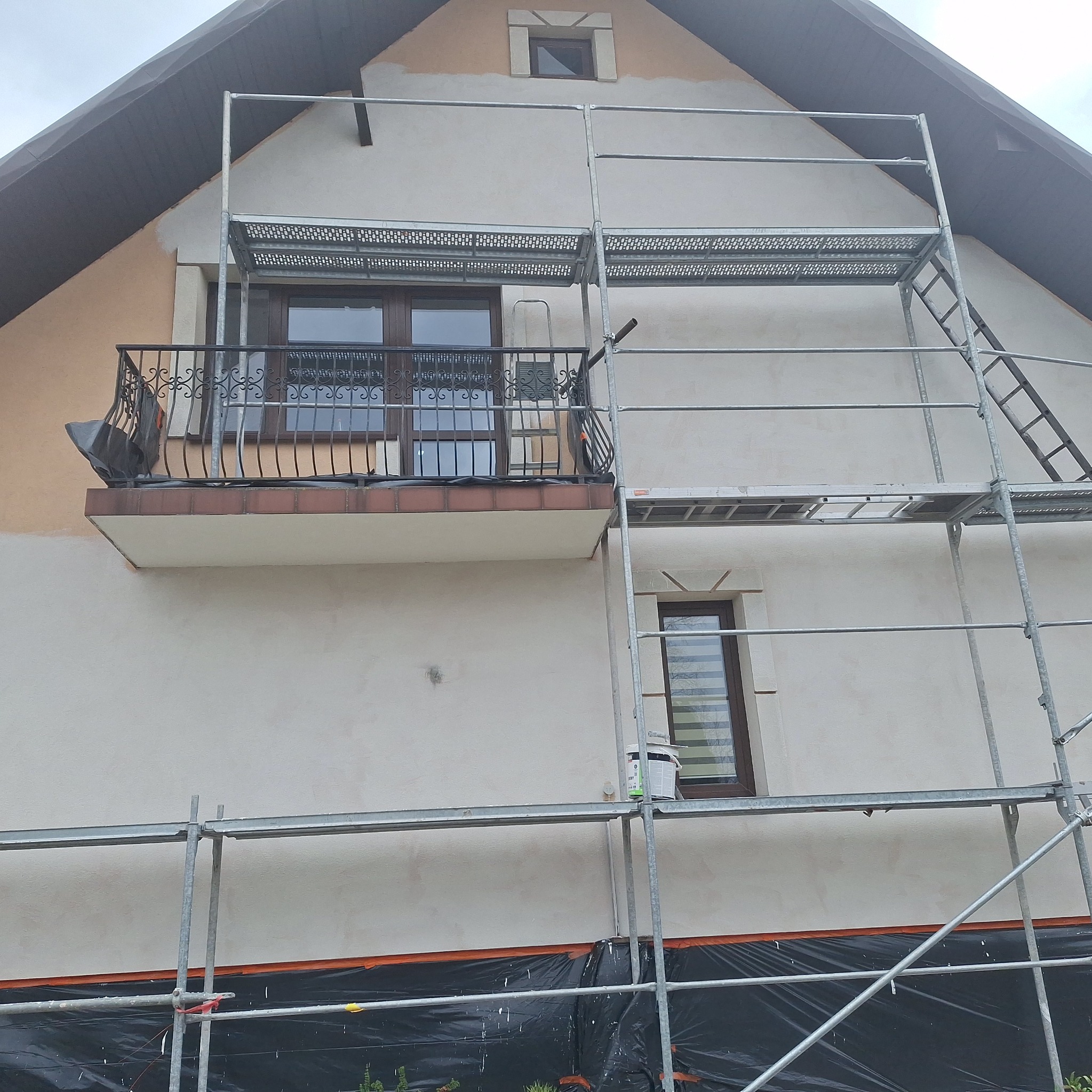 Dom w trakcie malowania elewacji, widoczne rusztowania, częściowo pomalowana ściana, zabezpieczone folią podłoże. Prace w toku, jasna kolorystyka.