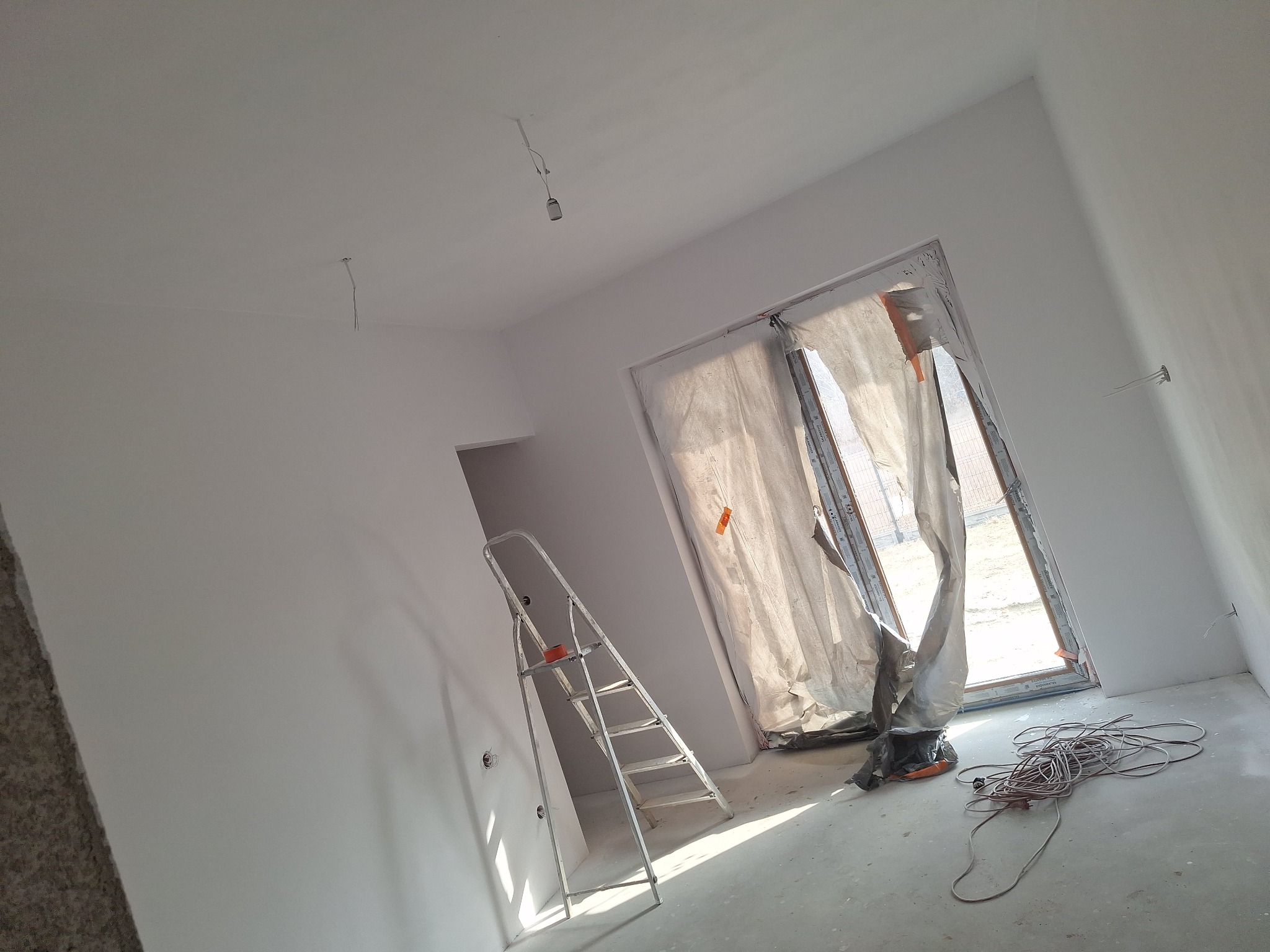 Wnętrze po malowaniu na biało, widoczna drabina, okno zabezpieczone folią malarską i zwój kabli na podłodze. Świeżo pomalowane ściany w nowym mieszkaniu.