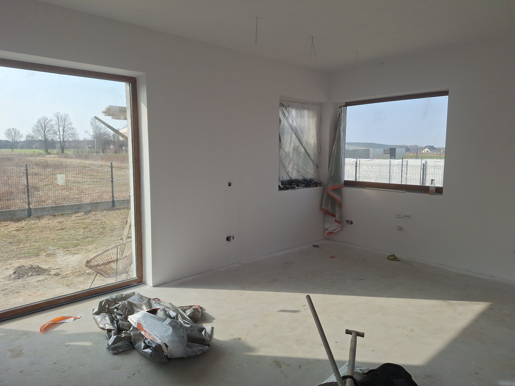 Wnętrze w trakcie prac wykończeniowych: świeżo pomalowane ściany na biało, okna zabezpieczone folią, widoczne narzędzia i materiały budowlane na podłodze.