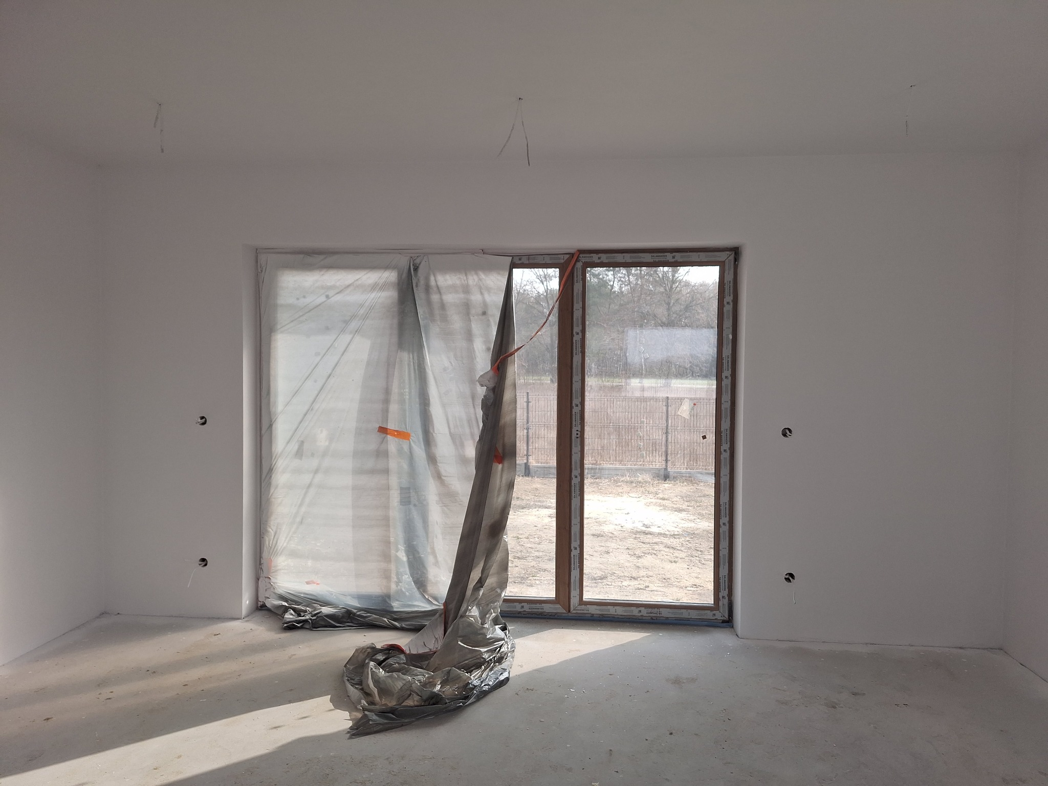 Wnętrze w stanie deweloperskim z oknem balkonowym zabezpieczonym folią malarską. Świeżo pomalowane ściany, widoczne wystające kable. Podłoga surowa, betonowa.