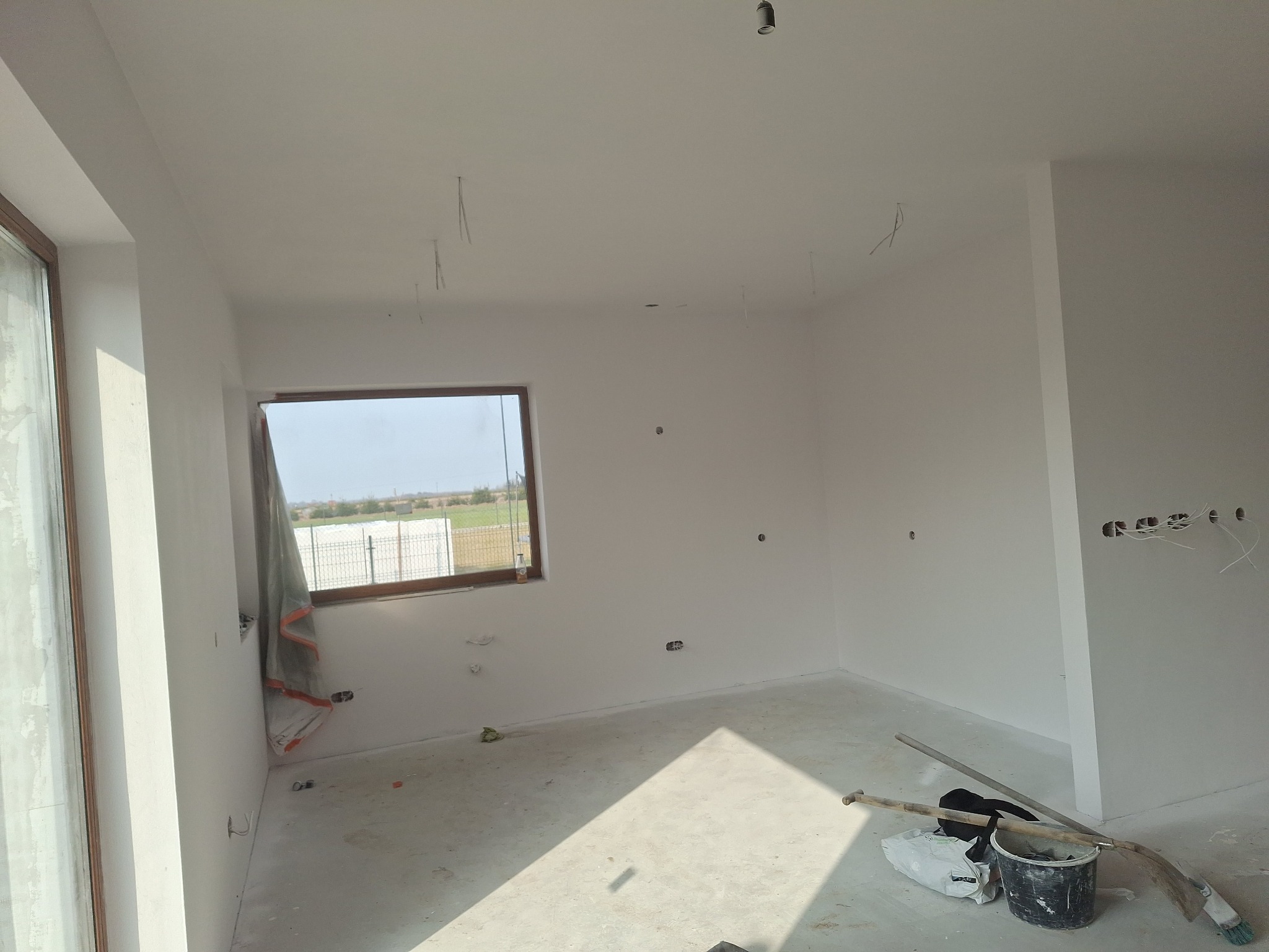 Białe, świeżo pomalowane wnętrze pokoju z oknem. Widoczne narzędzia malarskie i kable wystające ze ściany. Jasne, naturalne światło. Minimalistyczny styl wykończenia.