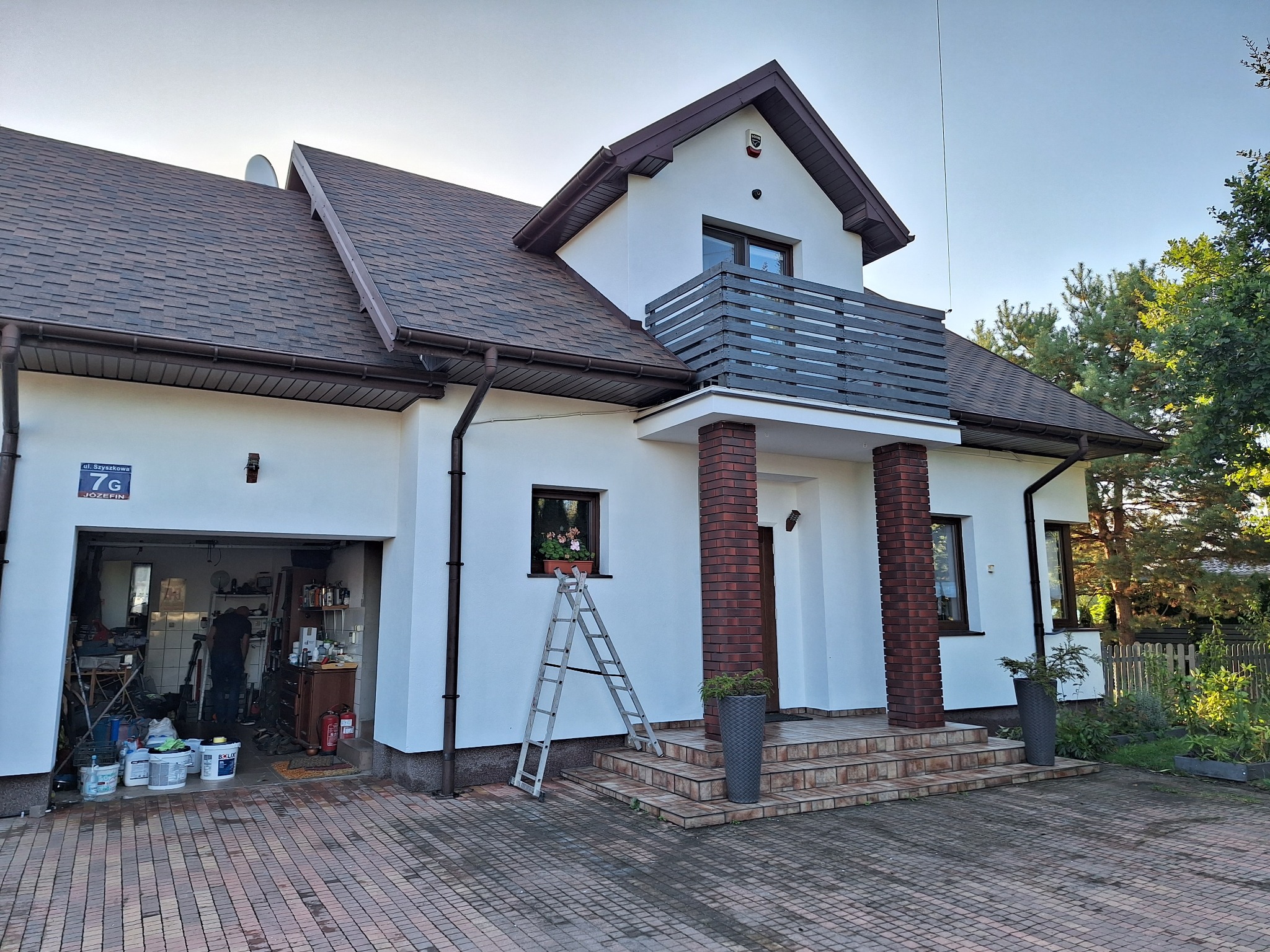 Biały dom z brązowym dachem po renowacji. Widoczna drabina oparta o ścianę, otwarty garaż z narzędziami i farbami. Świeżo pomalowana elewacja w słoneczny dzień.