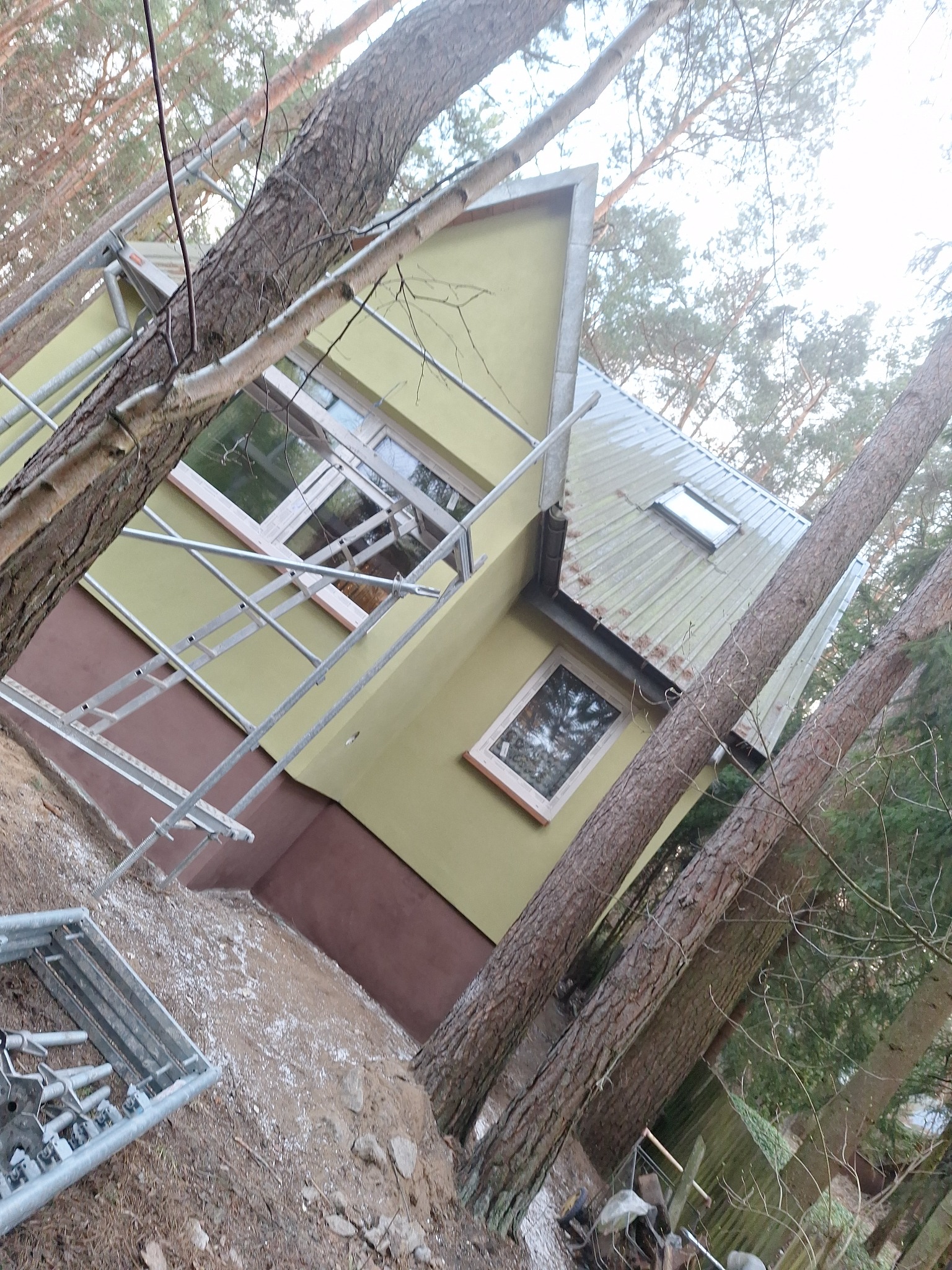 Malowanie elewacji domu w lesie, widoczne rusztowania i pomalowana dolna część budynku na brązowo, a górna na zielono. Ujęcie z dołu, drzewa na pierwszym planie.