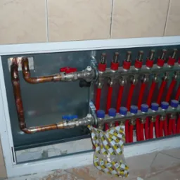 Rozdzielacz ogrzewania podłogowego w metalowej szafce, z miedzianymi rurami i czerwonymi przewodami, widoczny fragment opakowania Viega.