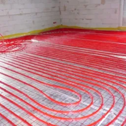 Instalacja ogrzewania podłogowego z czerwonych rur na folii odblaskowej z rastrem, widok na surową ścianę z betonu komórkowego i okno.