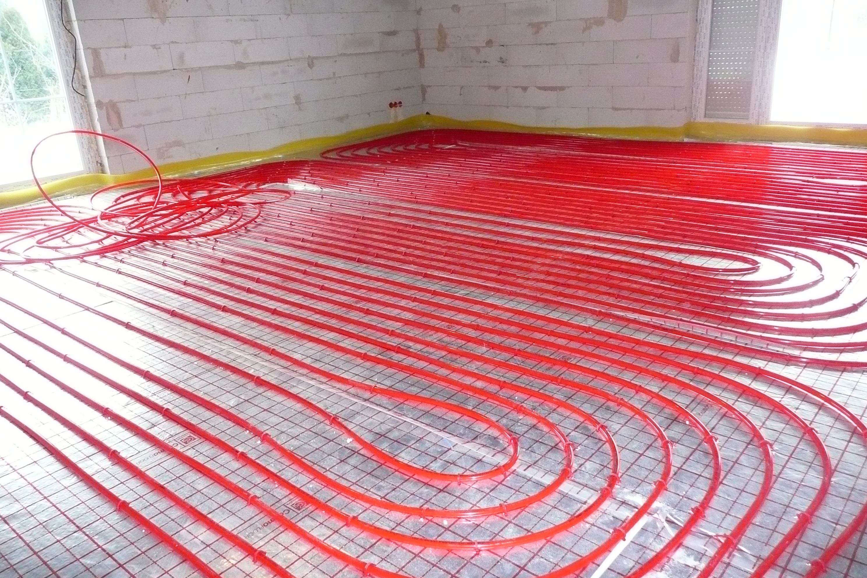 Instalacja ogrzewania podłogowego z czerwonych rur na folii odblaskowej z rastrem, widok na surową ścianę z betonu komórkowego i okno.