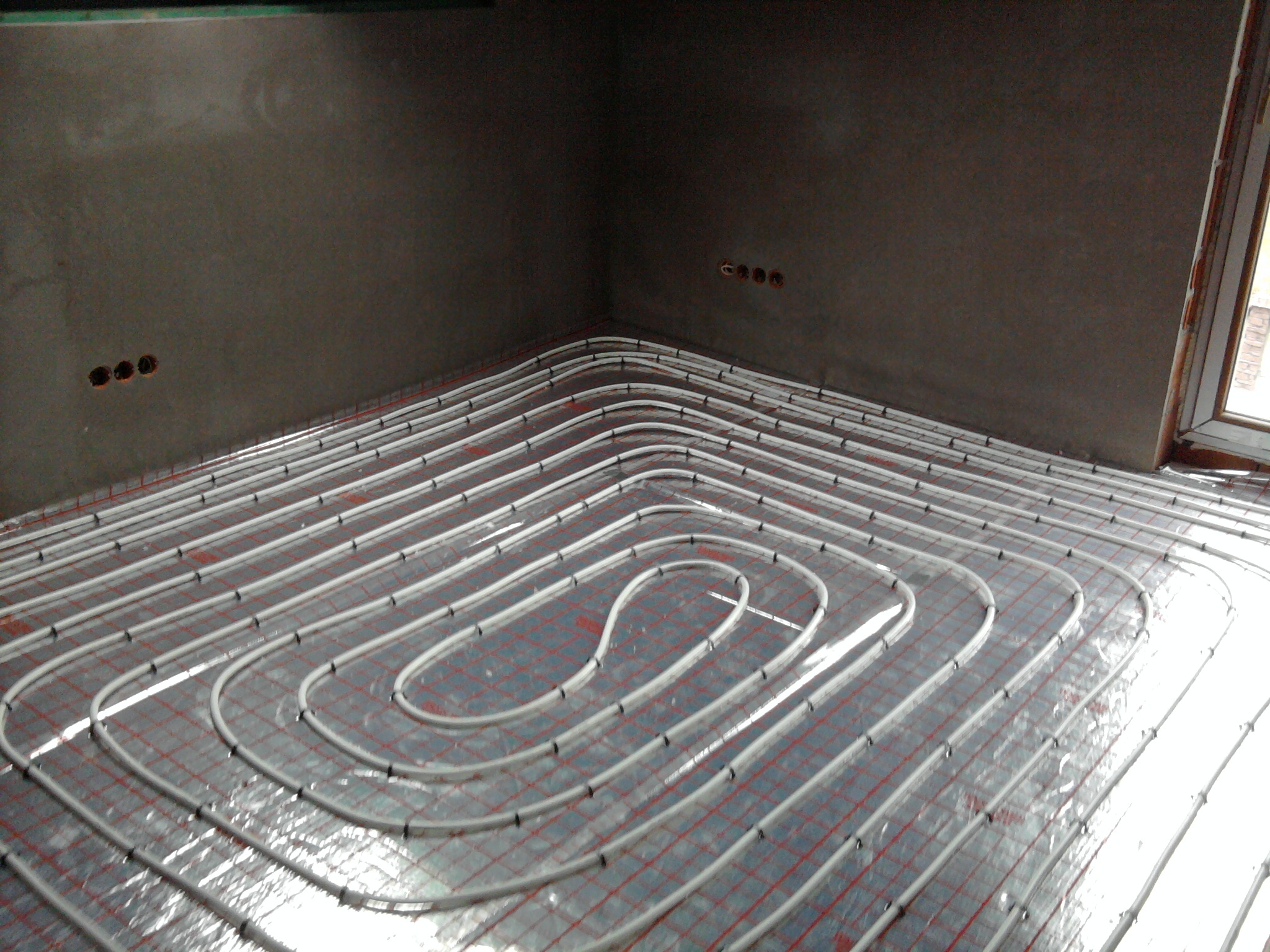 Szare, surowe wnętrze w trakcie instalacji ogrzewania podłogowego: białe rury ułożone na folii z czerwoną siatką, widoczne puszki elektryczne w ścianie i rama okienna.