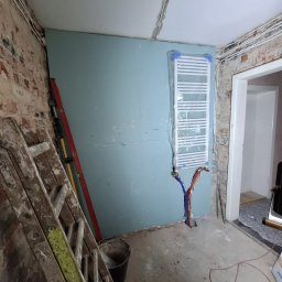 ENERGOHYDROINSTAL SPÓŁKA Z OGRANICZONĄ ODPOWIEDZIALNOŚCIĄ - Łazienka w trakcie remontu: grzejnik drabinkowy zabezpieczony folią, rury doprowadzone do ściany z płyt gipsowych, stara cegła, drabina malarska i poziomica.