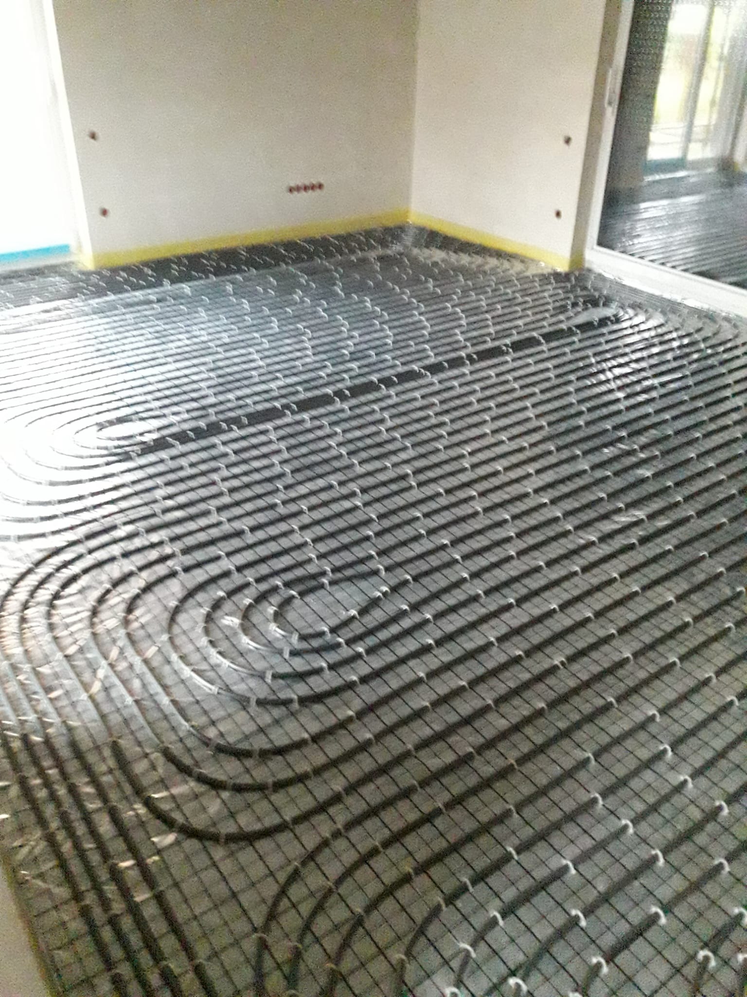 Ułożona instalacja ogrzewania podłogowego w jasnym pomieszczeniu, zabezpieczona taśmą przy ścianach. Widoczne pętle rur i elementy mocujące na folii izolacyjnej.