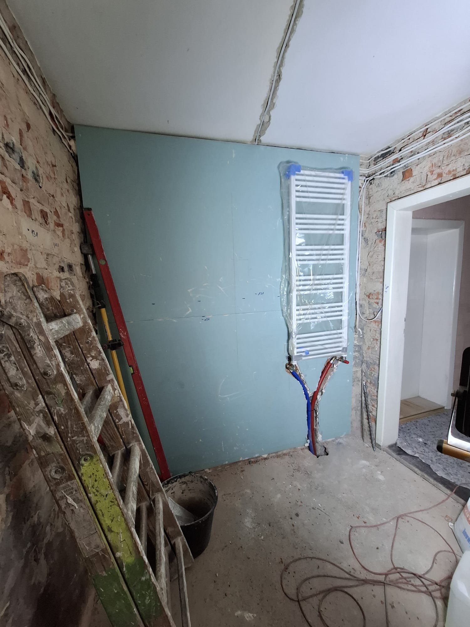 Łazienka w trakcie remontu: grzejnik drabinkowy zabezpieczony folią, rury doprowadzone do ściany z płyt gipsowych, stara cegła, drabina malarska i poziomica.