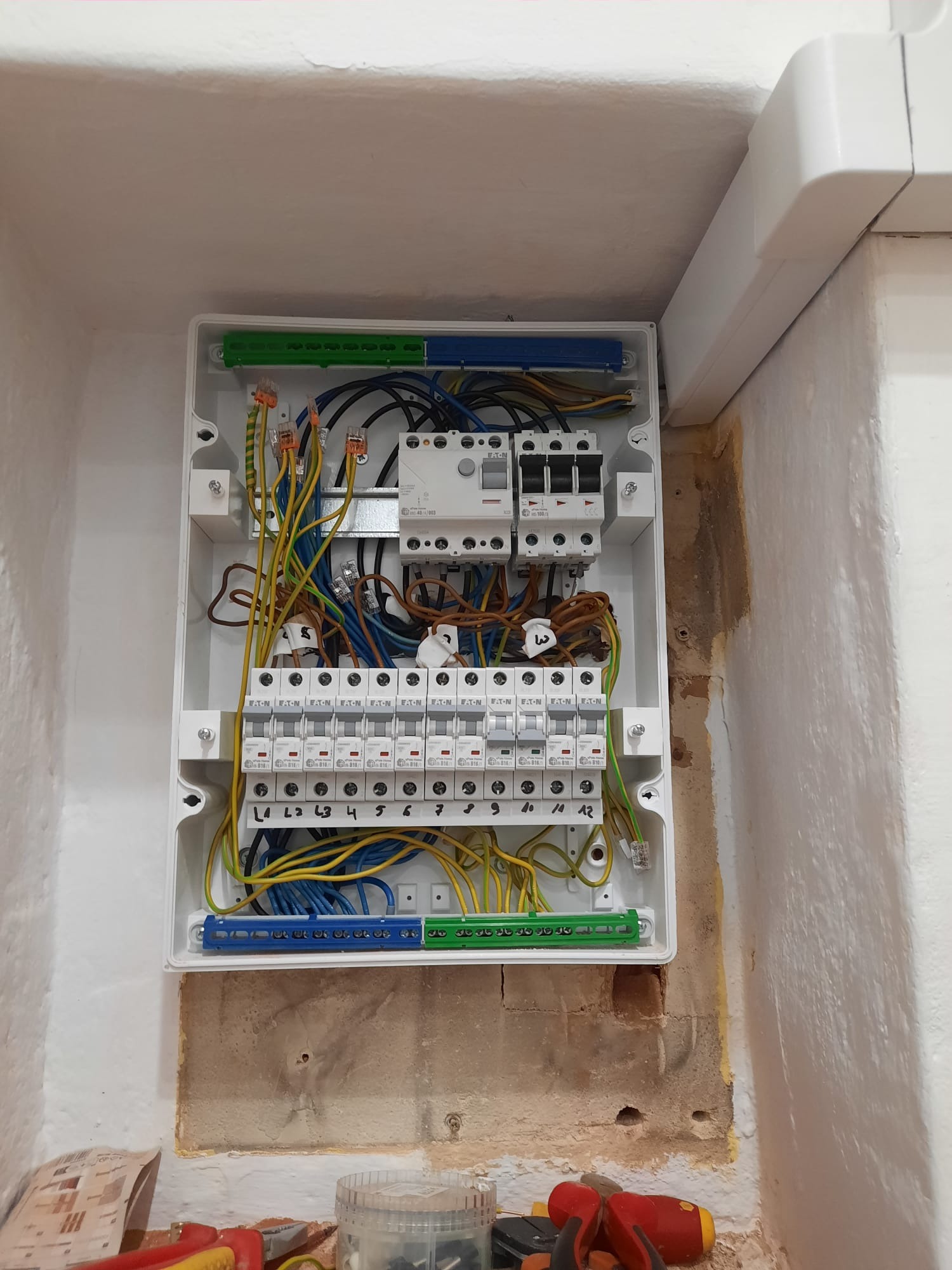 Otwarta skrzynka elektryczna z bezpiecznikami i przewodami wewnątrz, zamontowana na nierównej ścianie. Widoczne narzędzia u dołu kadru.