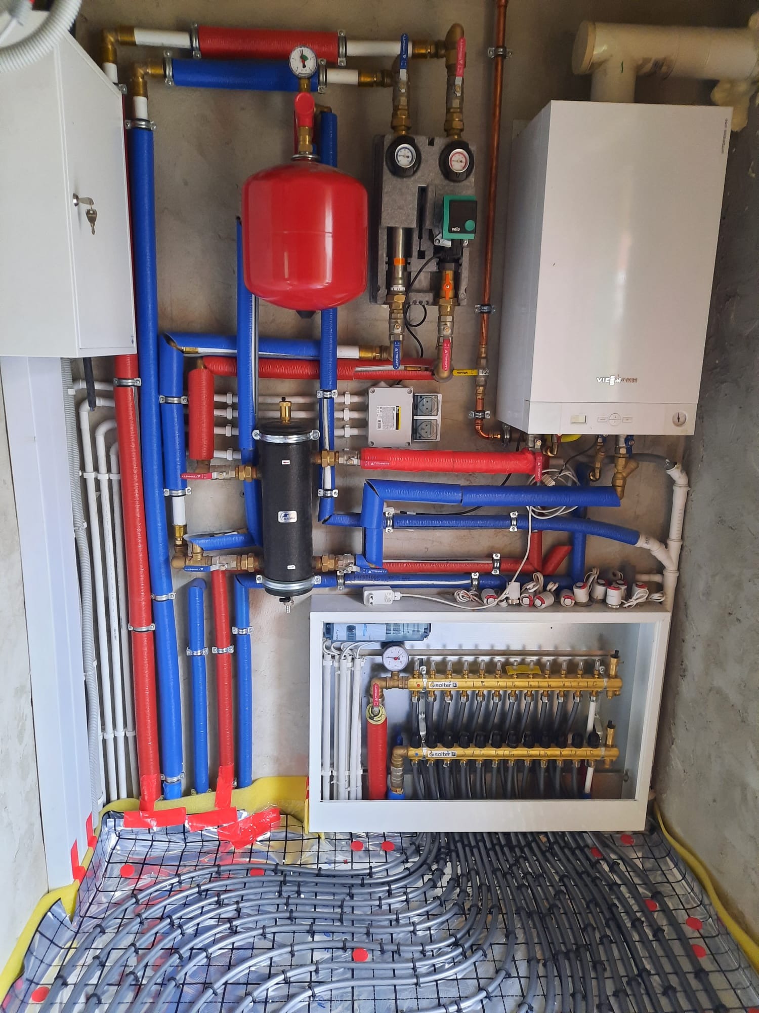 Kompleksowa instalacja grzewcza z piecem Viessmann, rozdzielaczem Solter i rurami w izolacji czerwono-niebieskiej. Widoczny system ogrzewania podłogowego przed wylewką.