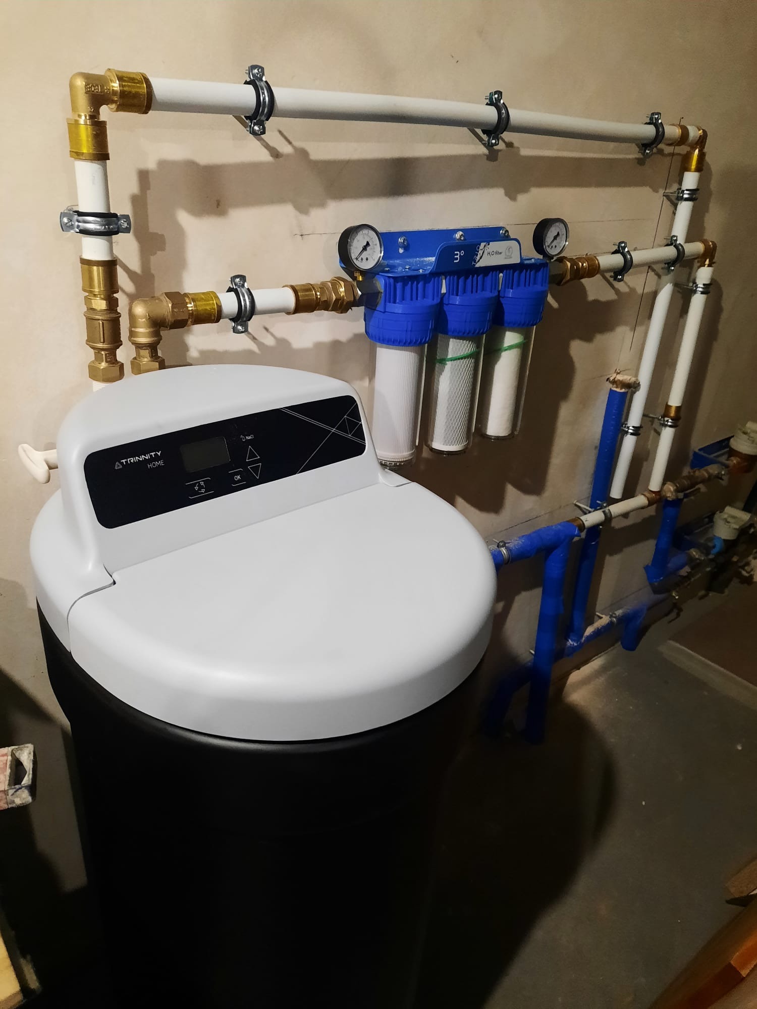 Zmontowany system uzdatniania wody Trinity Home z filtrami i manometrami, połączony rurami z mosiężnymi złączkami, zamocowany do ściany. Widoczne niebieskie rury.