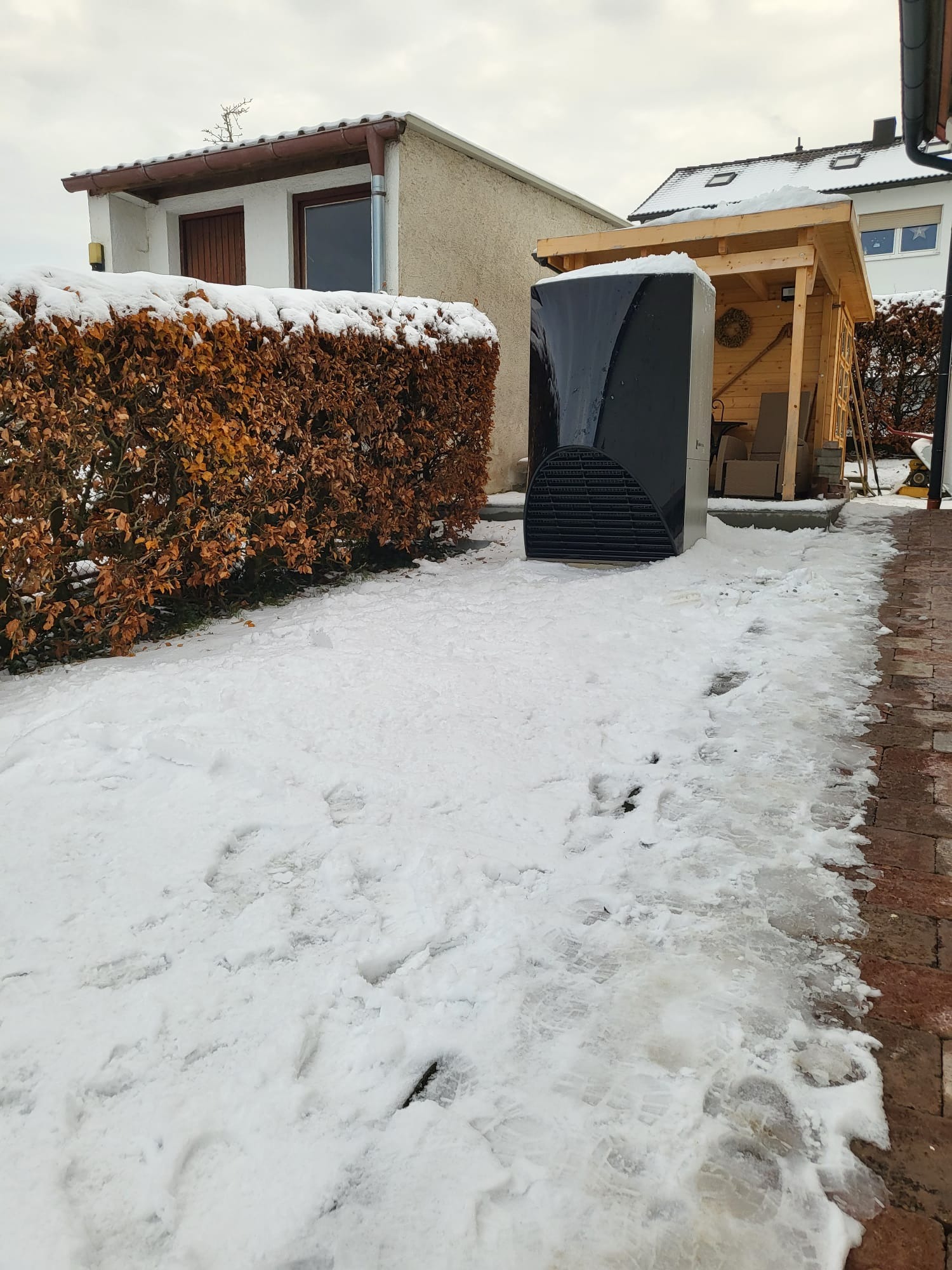 Czarna pompa ciepła powietrze-woda, zainstalowana na zewnątrz budynku w zimowej scenerii, obok drewnianej altany i żywopłotu. Ślady na śniegu.