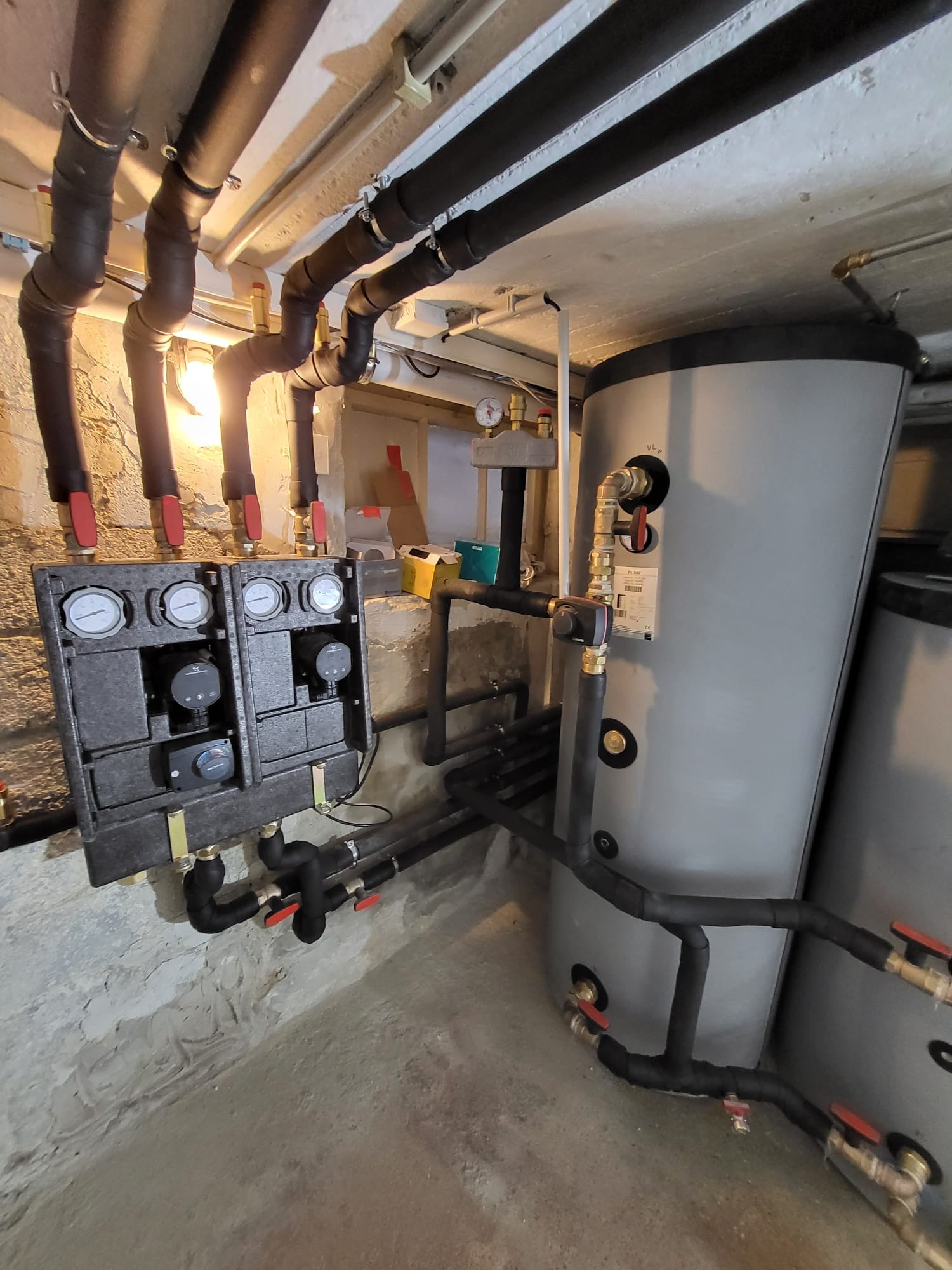 Szara instalacja grzewcza z czarnymi rurami i zaworami z czerwonymi uchwytami, zamontowana w piwnicy z surowymi ścianami. Widoczne manometry i zbiornik.