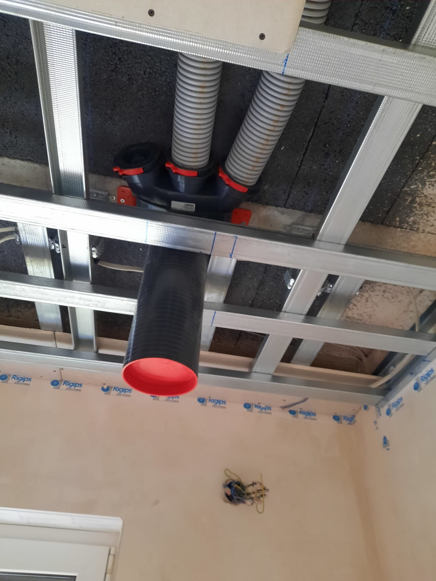 Szara rura wentylacyjna z czerwonymi elementami, zamontowana pod sufitem podwieszanym z profili Rigips, widoczne przewody elektryczne w ścianie.