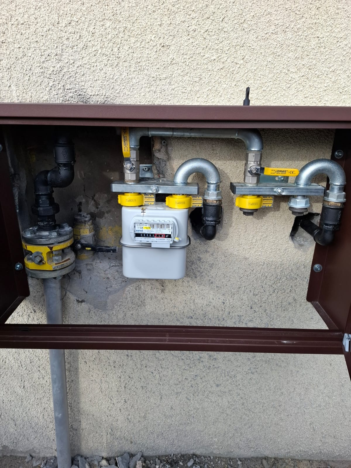 Zamknięta skrzynka z gazomierzem i instalacją gazową na zewnątrz budynku. Widoczne rury, zawory i elementy mocujące. Szara ściana z fakturą w tle.