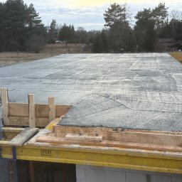 DMK Budownictwo Dominik Małecki - Wylany fundament pod budowę domu jednorodzinnego. Widoczne szalunki PERI, zbrojenie i świeży beton. W tle drzewa i częściowo widoczny budynek.