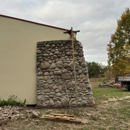 DMK Budownictwo Dominik Małecki - Fragment elewacji budynku z kamienia polnego, z widocznym rusztowaniem pomocniczym. Tło stanowi ściana budynku o jasnej elewacji i jesienne otoczenie.