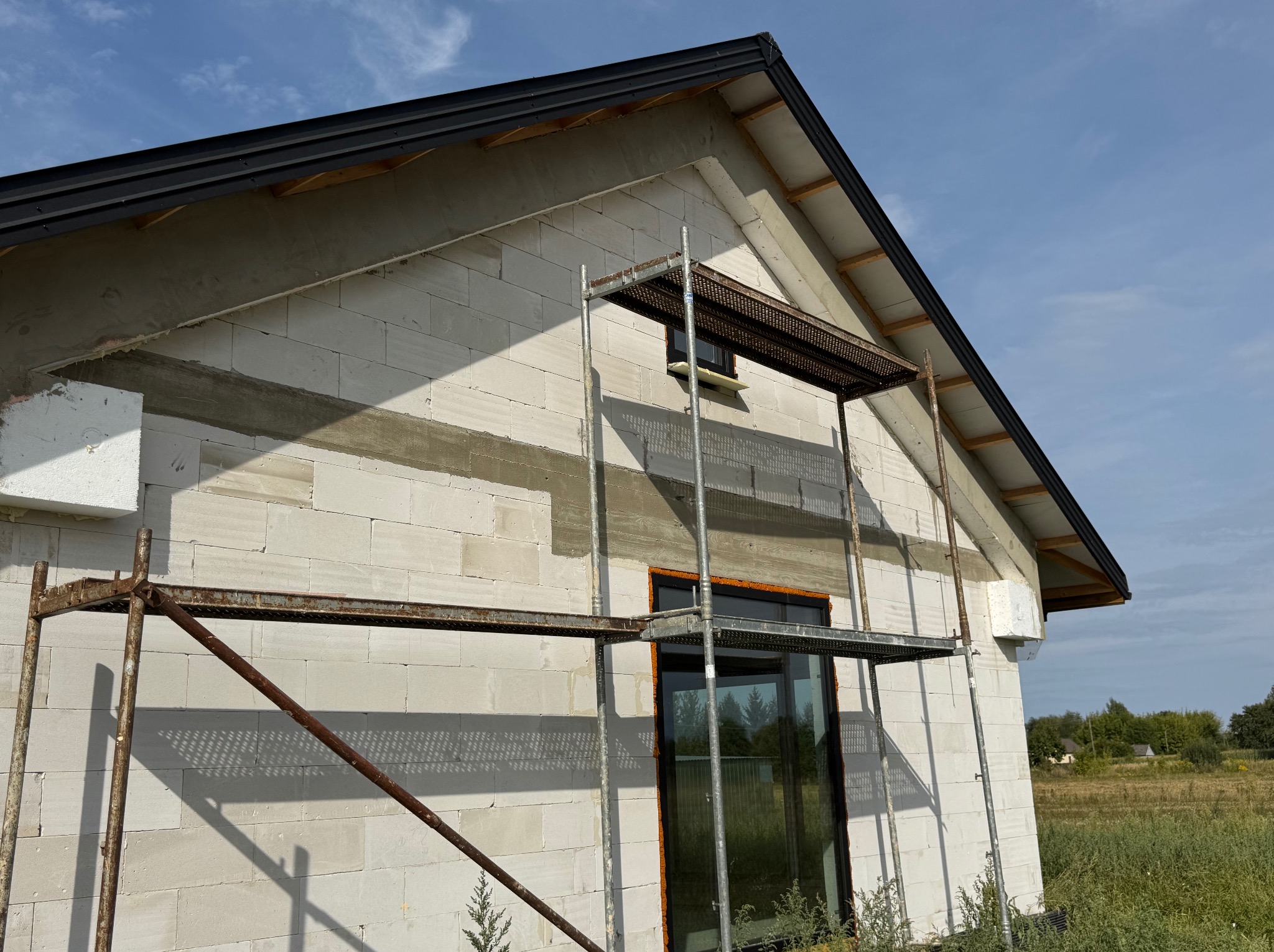 Budowa domu z białych bloczków, widoczne rusztowania i okna. Częściowo ocieplona ściana szczytowa. Tło: łąka i niebo z chmurami.