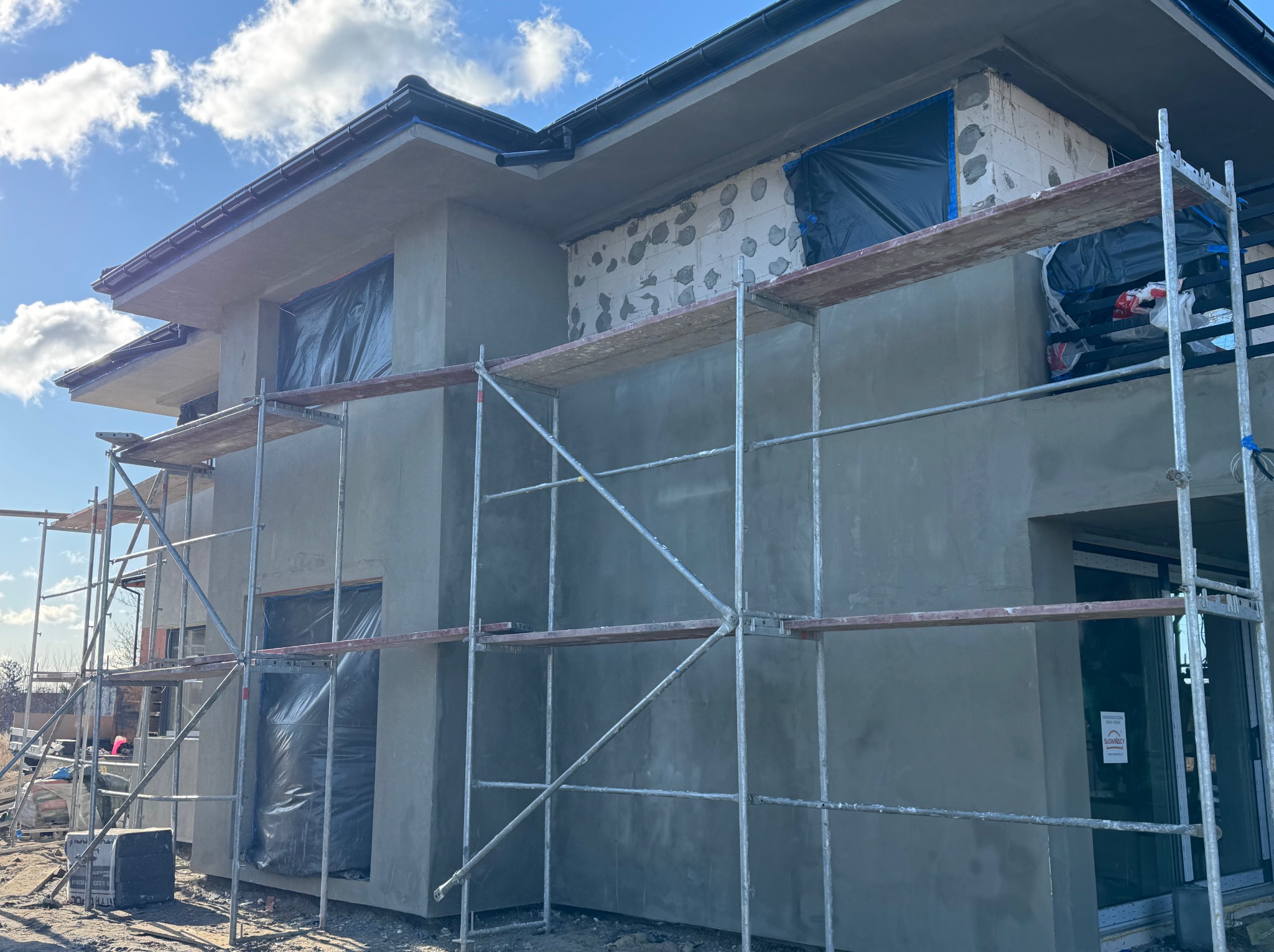 Ocieplony dom w trakcie prac elewacyjnych, widoczne rusztowania i okna zabezpieczone folią. Świeżo nałożony tynk w kolorze szarym. Częściowo otynkowana ściana z pustaków.