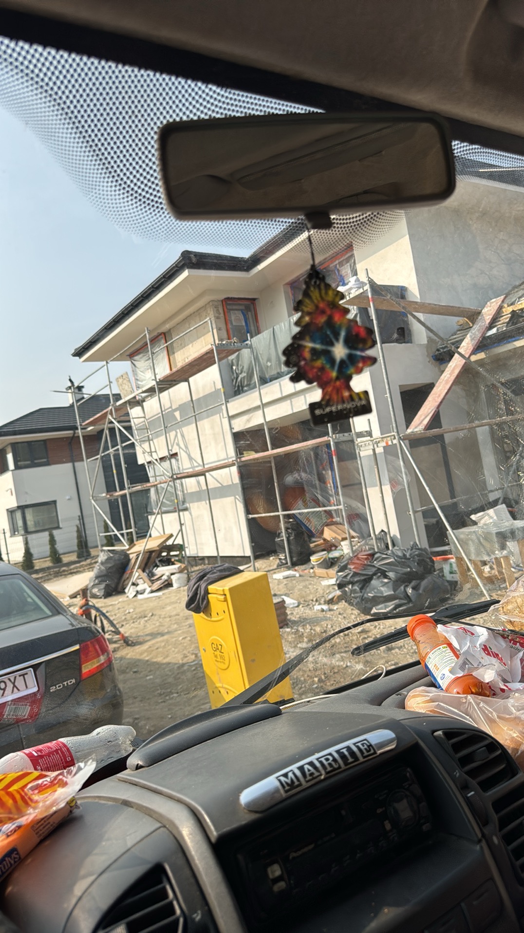 Dom w trakcie ocieplania elewacji, widok z wnętrza samochodu. Na pierwszym planie deska rozdzielcza, w tle rusztowania i fragmenty ocieplonej ściany.