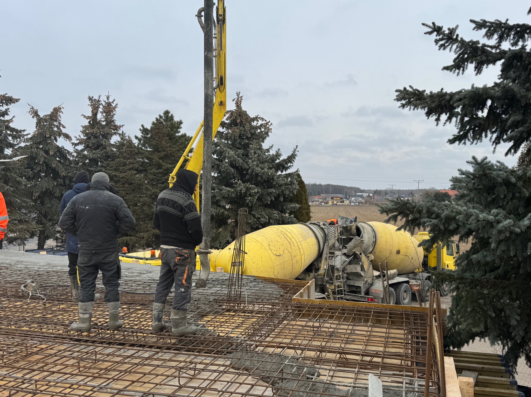 Wylewanie betonu na zbrojenie stropu z użyciem pompy do betonu. Żółta betoniarka w tle, pracownicy w roboczych ubraniach. Widok na drzewa i zabudowania w oddali.