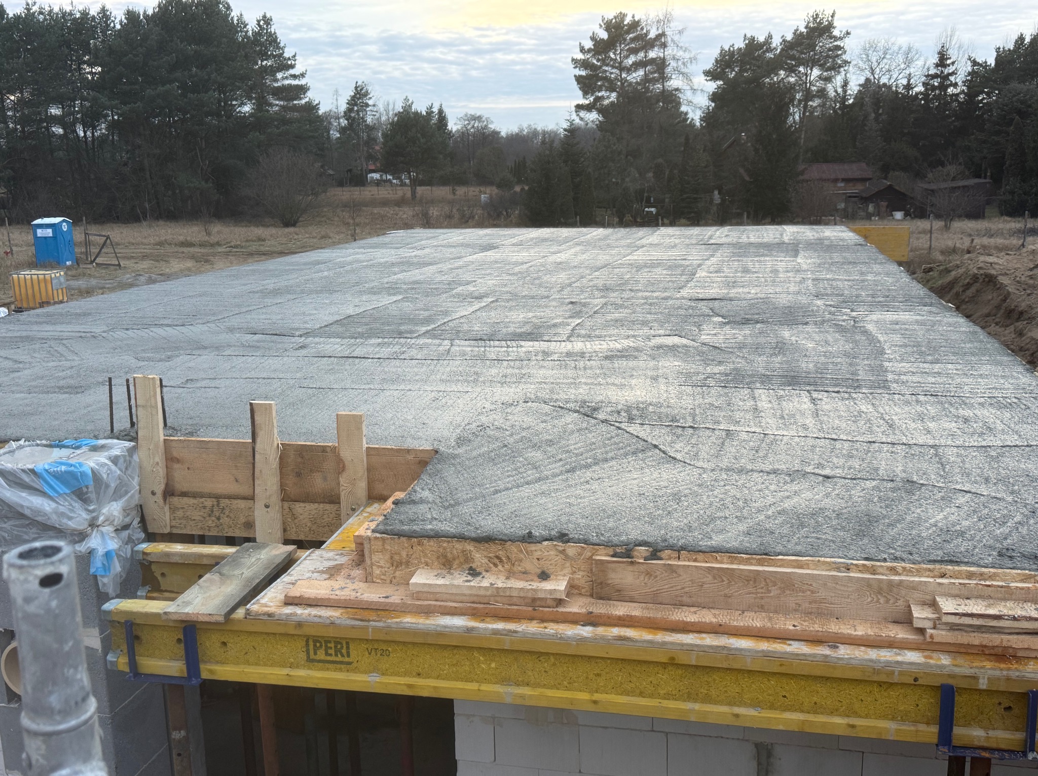 Wylany fundament pod budowę domu jednorodzinnego. Widoczne szalunki PERI, zbrojenie i świeży beton. W tle drzewa i częściowo widoczny budynek.
