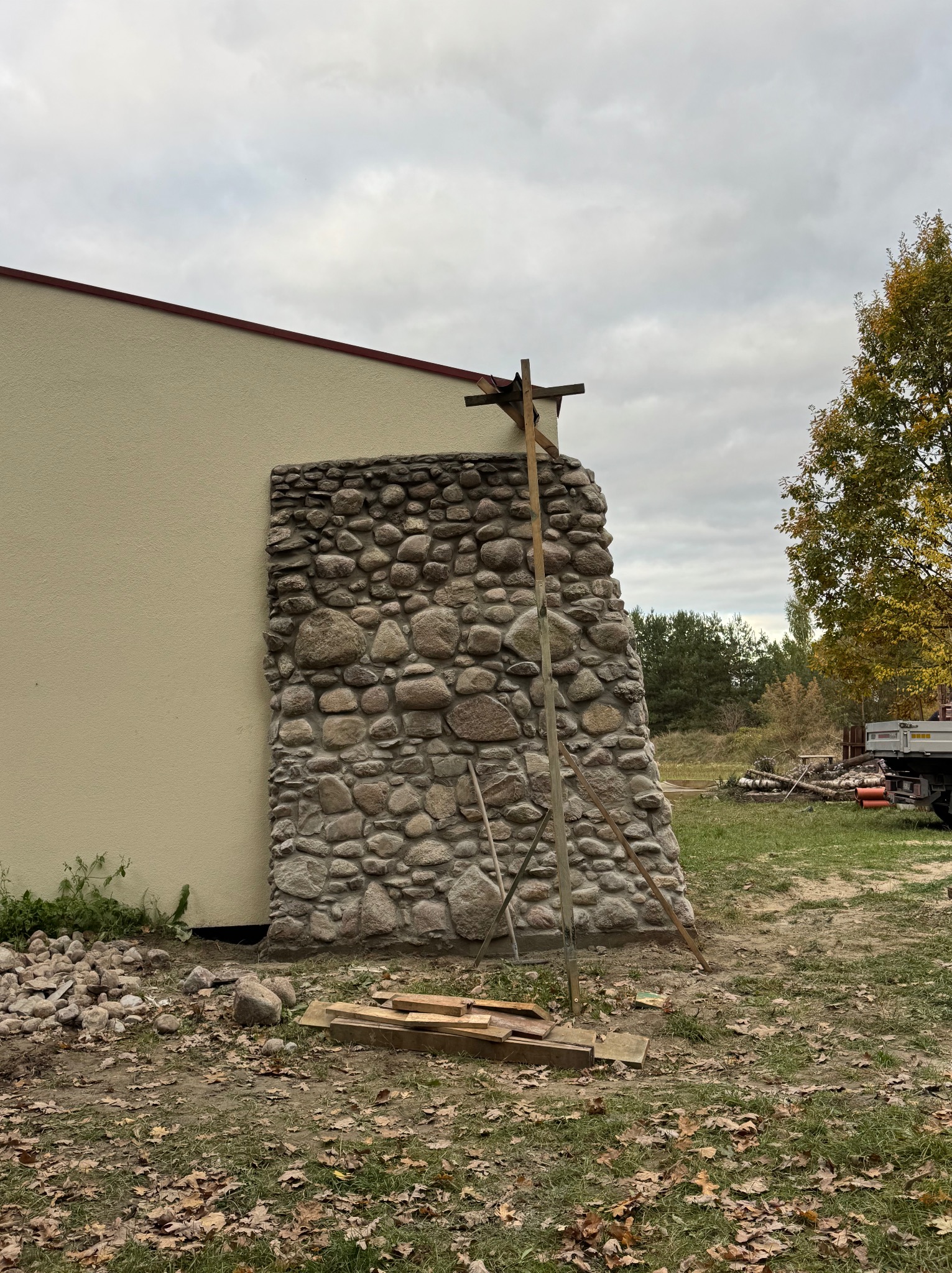 Fragment elewacji budynku z kamienia polnego, z widocznym rusztowaniem pomocniczym. Tło stanowi ściana budynku o jasnej elewacji i jesienne otoczenie.