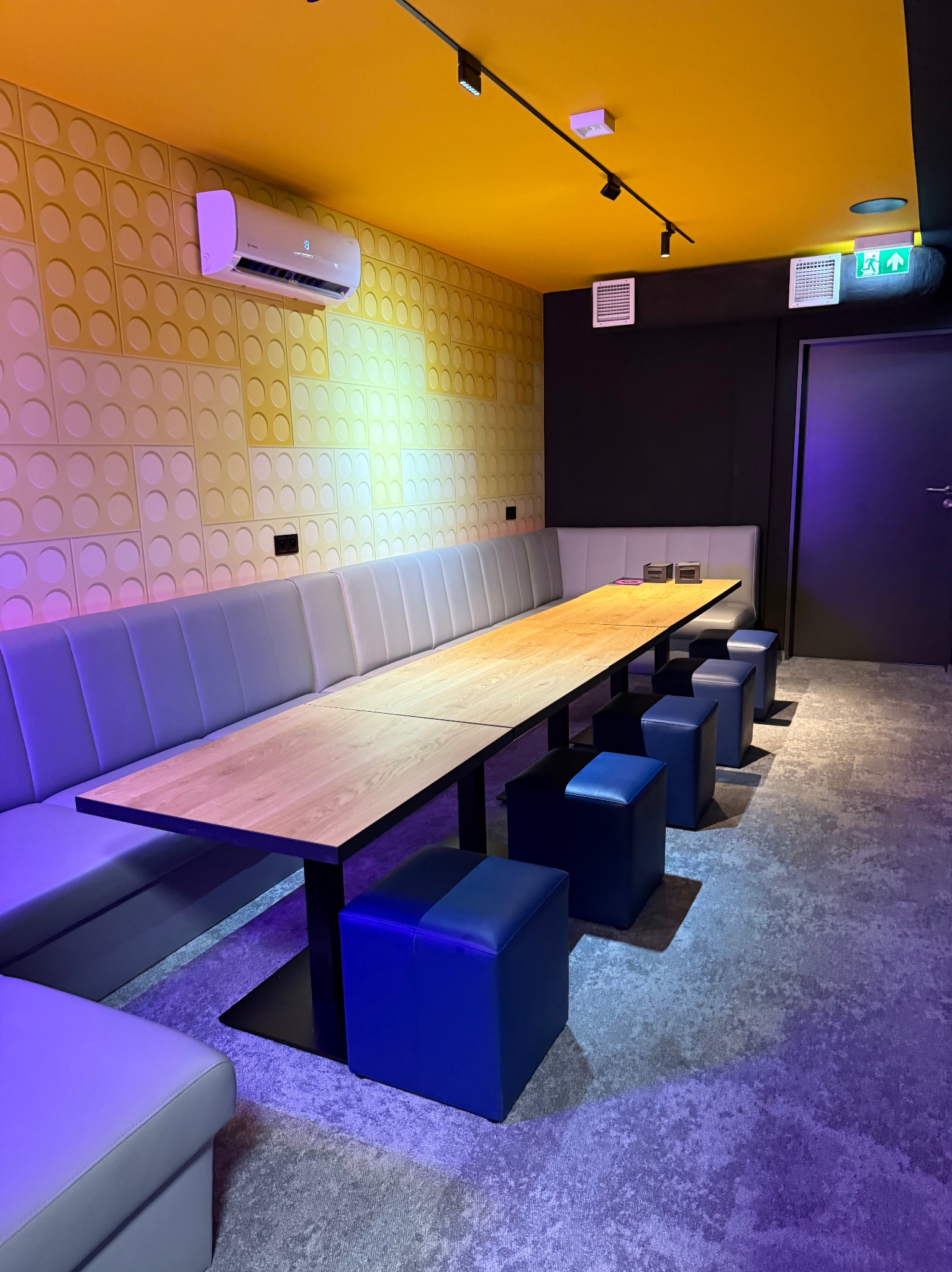 Nowoczesne wnętrze: długa drewniana ława z tapicerowanymi siedziskami, żółty sufit, designerska ściana z fakturą i klimatyzacja. Oświetlenie punktowe.