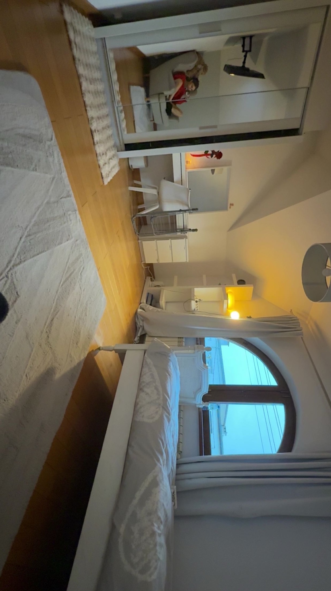 Jasna sypialnia z drewnianą podłogą, białymi meblami i oknem w kształcie łuku. W lustrzanej szafie odbija się osoba siedząca w fotelu. Ciepłe światło lampki nocnej.