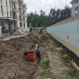 HYDROTECH Tomasz Woźniak - Pomarańczowa studzienka kanalizacyjna w wykopie ziemnym na tle budynków w trakcie budowy. Widoczne rury i izolacja fundamentów. Prace ziemne w toku.
