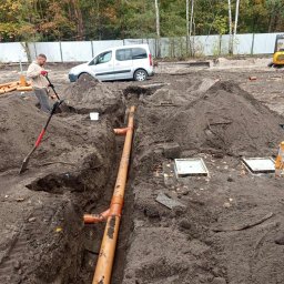 HYDROTECH Tomasz Woźniak - Montaż pomarańczowych rur kanalizacyjnych w wykopie, widoczny pracownik z łopatą, biały samochód i koparka w tle. Prace ziemne w toku.