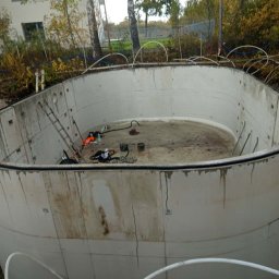 HYDROTECH Tomasz Woźniak - Betonowy zbiornik retencyjny w trakcie budowy, widoczne ślady po zalewaniu betonem, drabina i narzędzia wewnątrz. Tło stanowią drzewa i budynek.