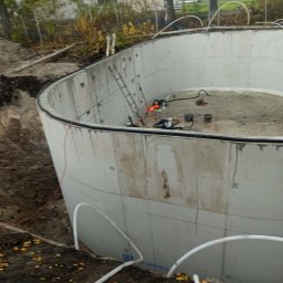 HYDROTECH Tomasz Woźniak - Betonowy zbiornik w ziemi, prawdopodobnie szambo lub basen w trakcie budowy. Widoczne rury, drabina i narzędzia. Otoczenie: ziemia, drzewa, budynek w tle.
