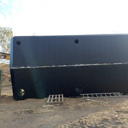 HYDROTECH Tomasz Woźniak - Dwa duże, czarne, betonowe zbiorniki z otworami, leżące na piasku. W tle widoczny żółty dźwig i drzewo. Na jednym ze zbiorników widnieje biała cyfra '3'.