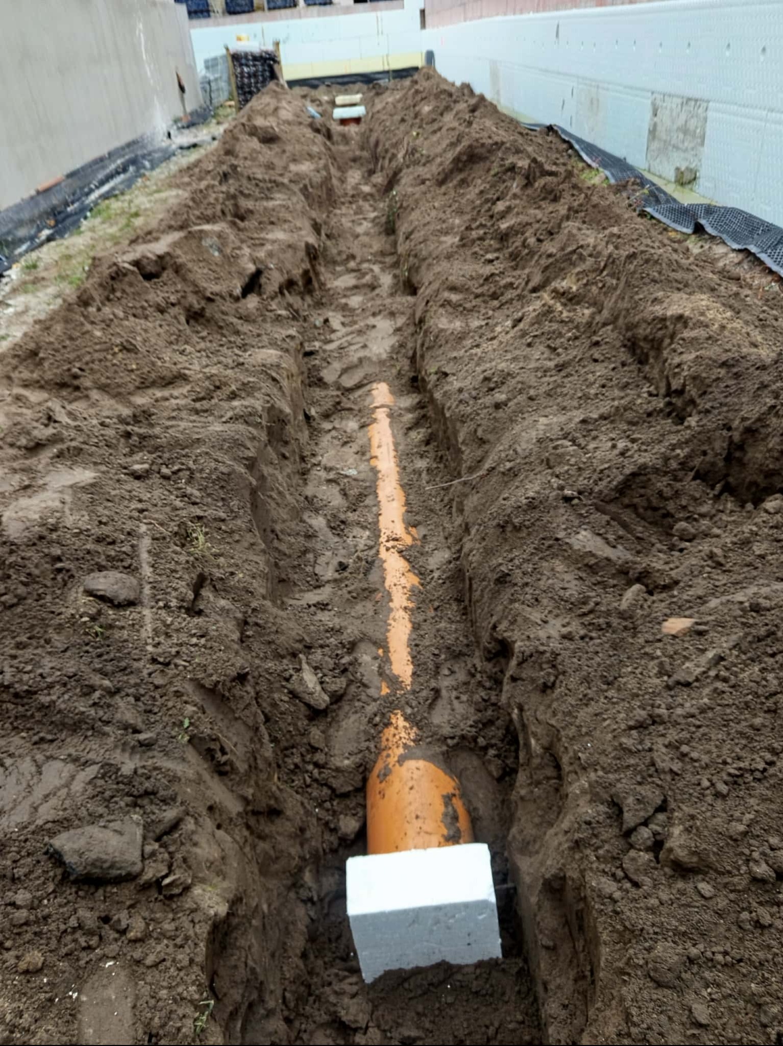 Pomarańczowa rura kanalizacyjna ułożona w wykopie ziemnym, zabezpieczona białym bloczkiem. Widok z góry na wykop wzdłuż ściany budynku z izolacją.