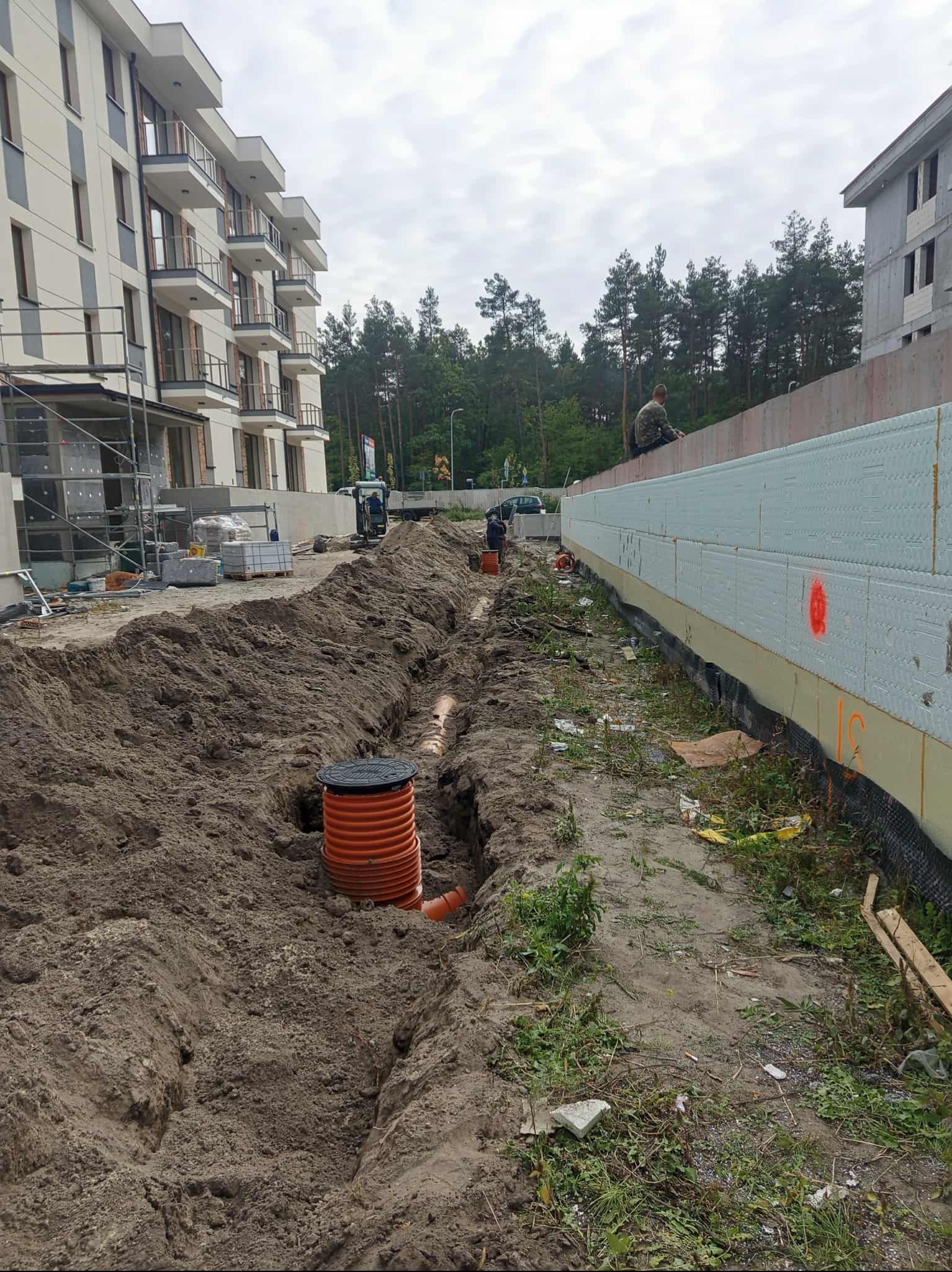 Pomarańczowa studzienka kanalizacyjna w wykopie ziemnym na tle budynków w trakcie budowy. Widoczne rury i izolacja fundamentów. Prace ziemne w toku.