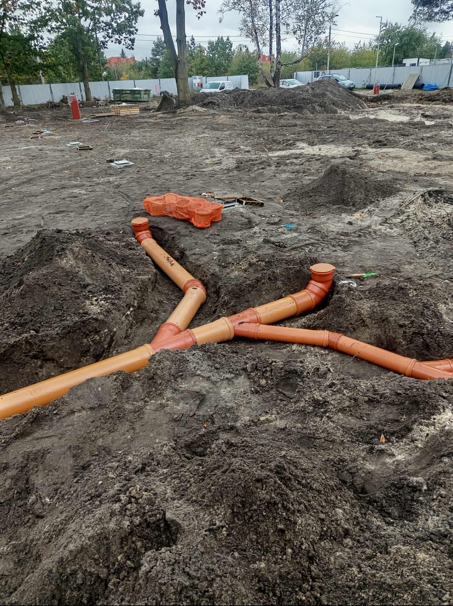 Pomarańczowe rury kanalizacyjne ułożone w wykopie ziemnym na placu budowy. Widoczne elementy montażowe i narzędzia. W tle ogrodzenie i zieleń.