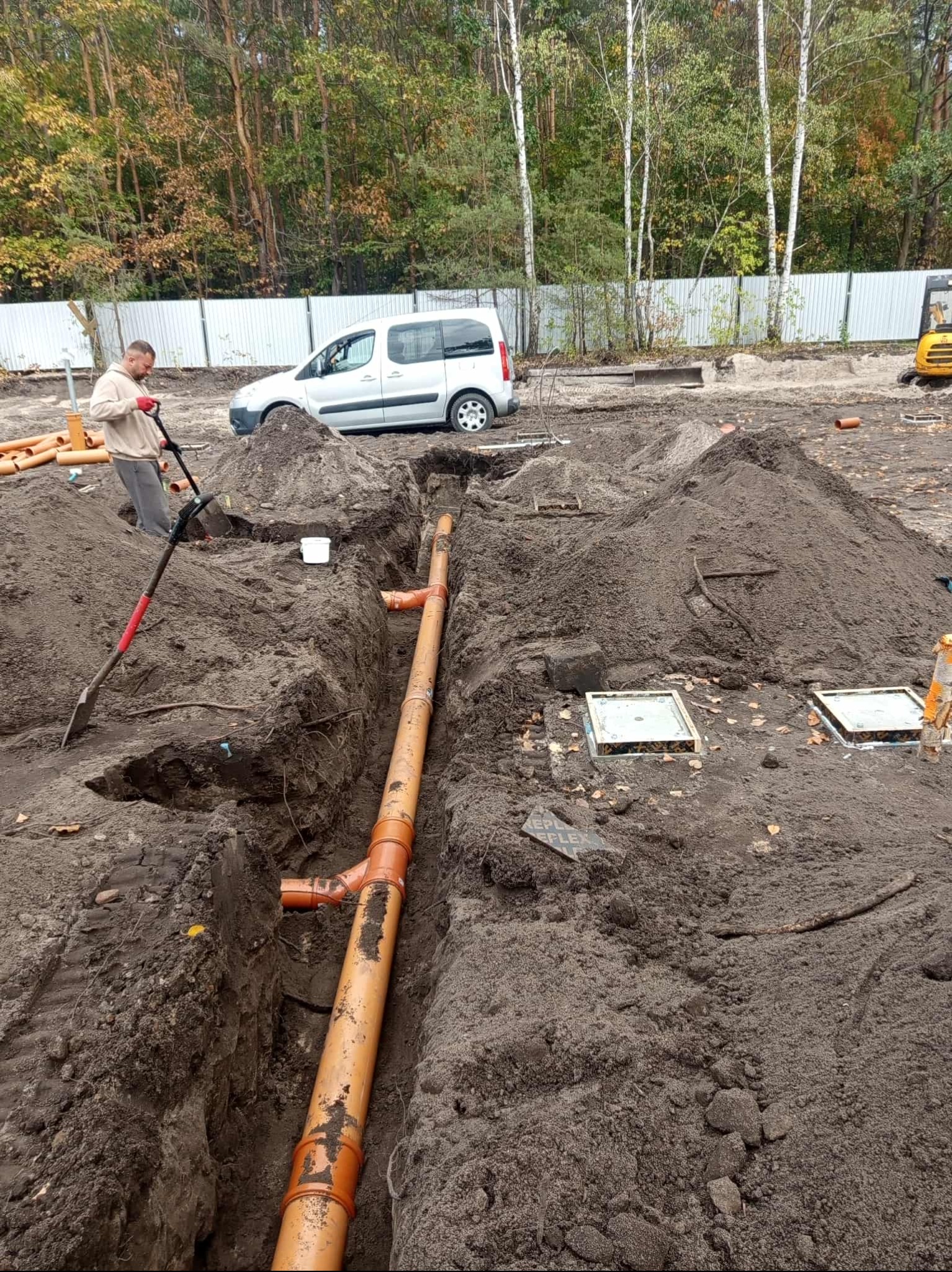 Montaż pomarańczowych rur kanalizacyjnych w wykopie, widoczny pracownik z łopatą, biały samochód i koparka w tle. Prace ziemne w toku.