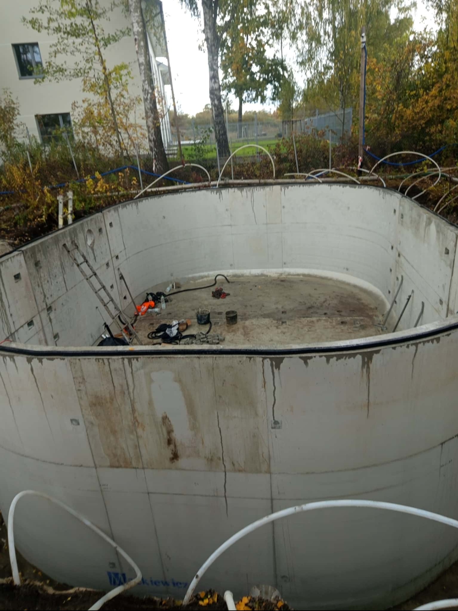Betonowy zbiornik retencyjny w trakcie budowy, widoczne ślady po zalewaniu betonem, drabina i narzędzia wewnątrz. Tło stanowią drzewa i budynek.