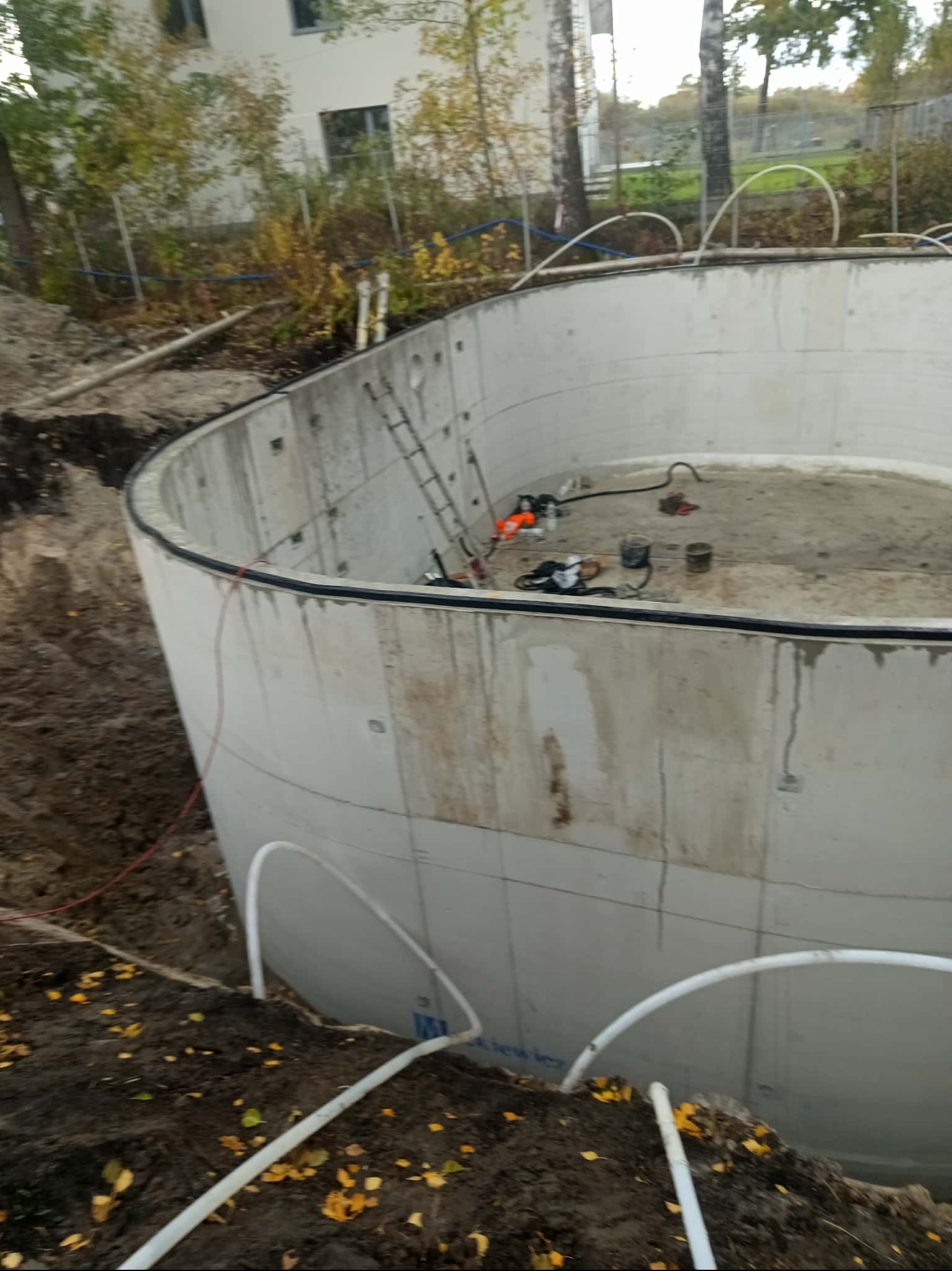 Betonowy zbiornik w ziemi, prawdopodobnie szambo lub basen w trakcie budowy. Widoczne rury, drabina i narzędzia. Otoczenie: ziemia, drzewa, budynek w tle.