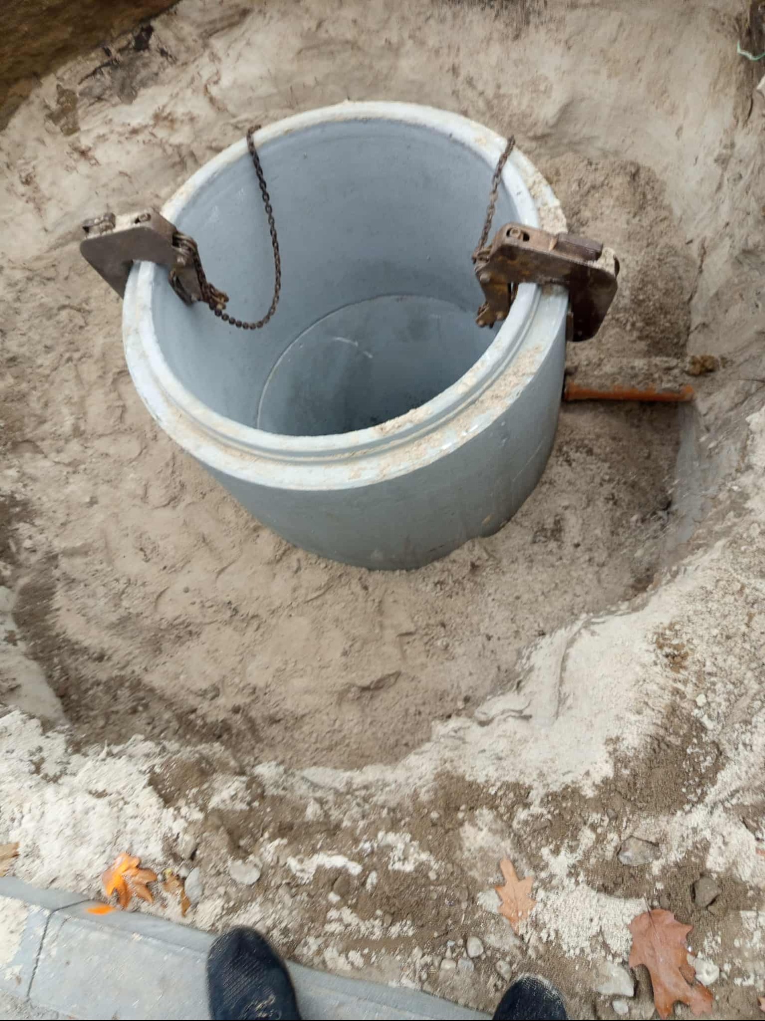 Betonowy krąg szamba w wykopie, podwieszony na łańcuchach z metalowymi uchwytami. Widoczny fragment rury i buty osoby fotografującej.