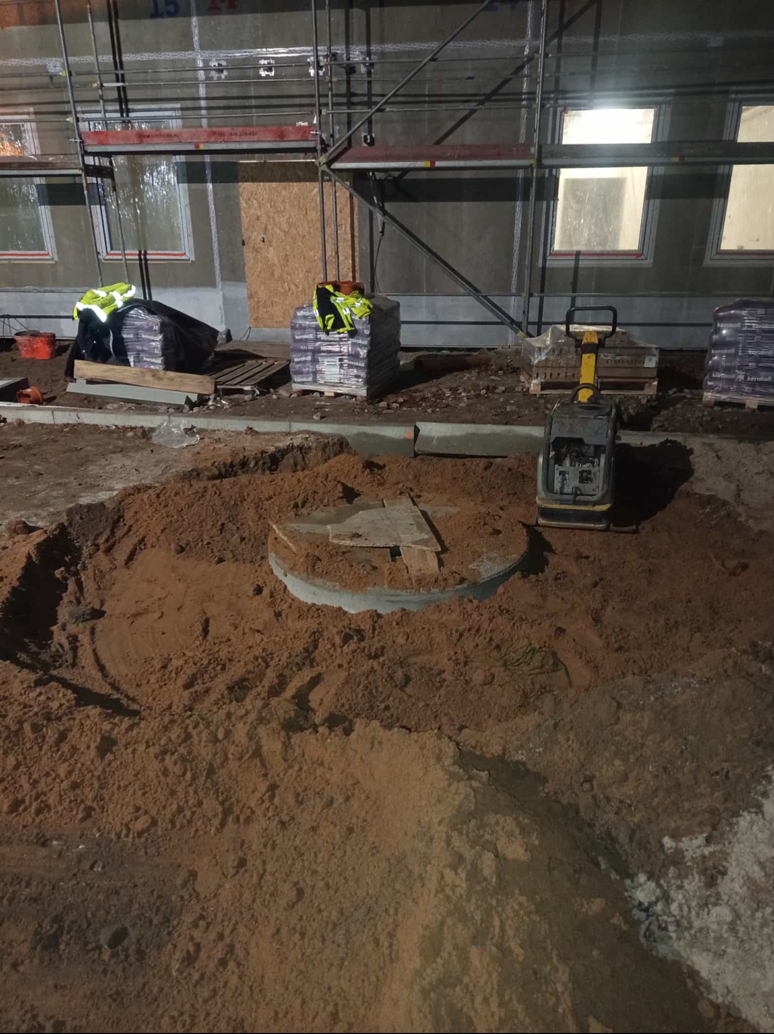 Wykonany wykop pod szambo betonowe z widocznym betonowym kręgiem. Na górze ubijarka, w tle materiały budowlane i rusztowanie. Praca w toku, nocne oświetlenie.