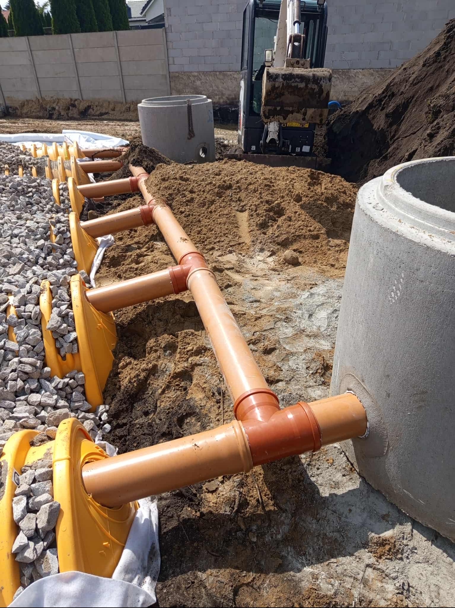 Montaż rur kanalizacyjnych z żółtymi osłonami w wykopie, widoczny betonowy krąg i koparka w tle. Etap budowy przyłącza kanalizacyjnego.