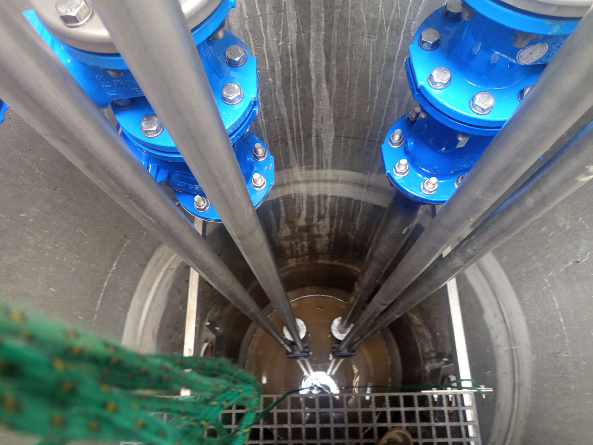 Wnętrze betonowego szamba z rurami i niebieskimi złączkami, widok z góry. W tle widoczne ślady po wodzie na ścianach zbiornika. Na dole kratka i fragment liny.